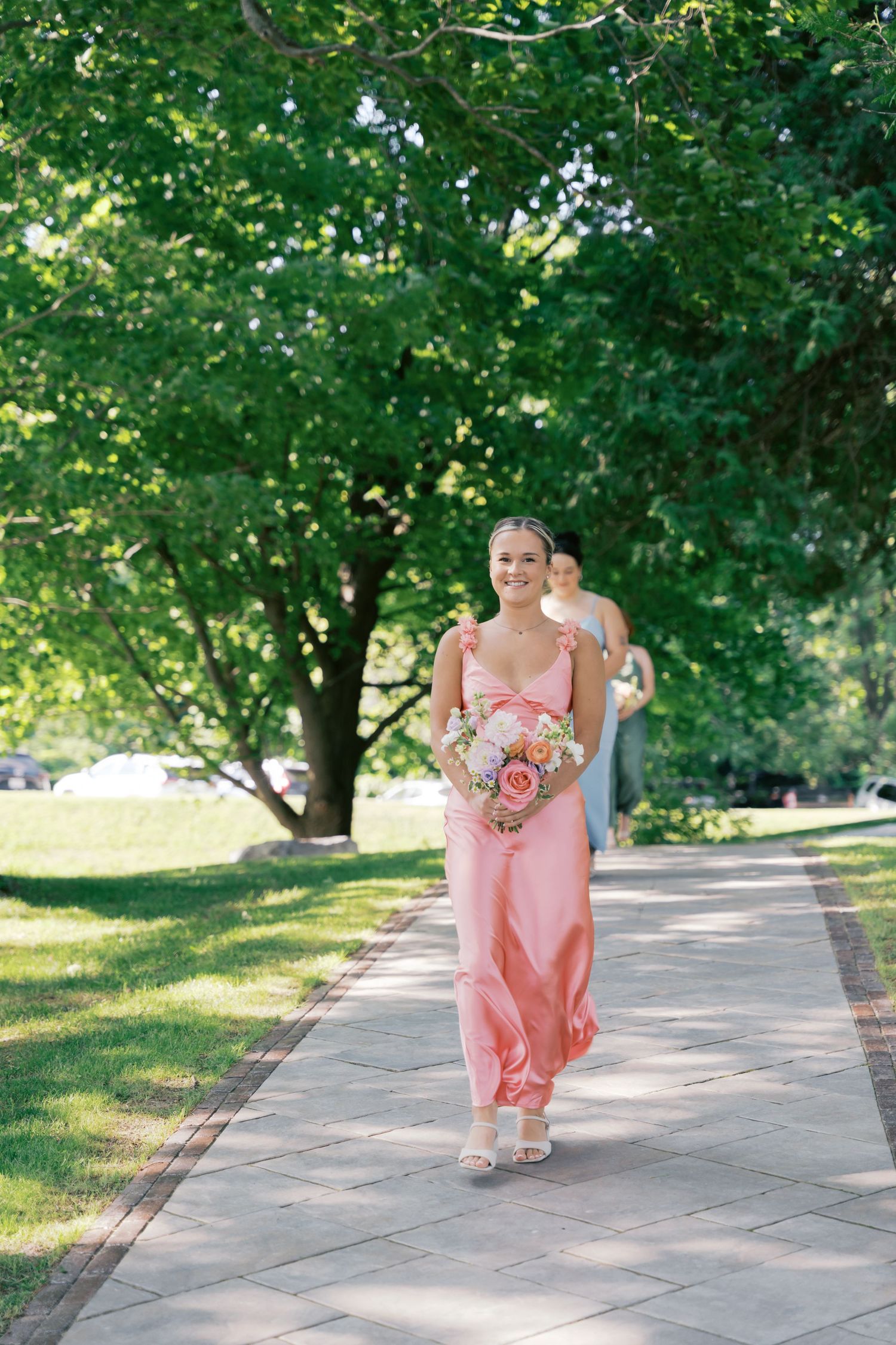 Someone in a flowing pink bridesmaid dress walks down a tree-lined path while holding a floral bouquet.