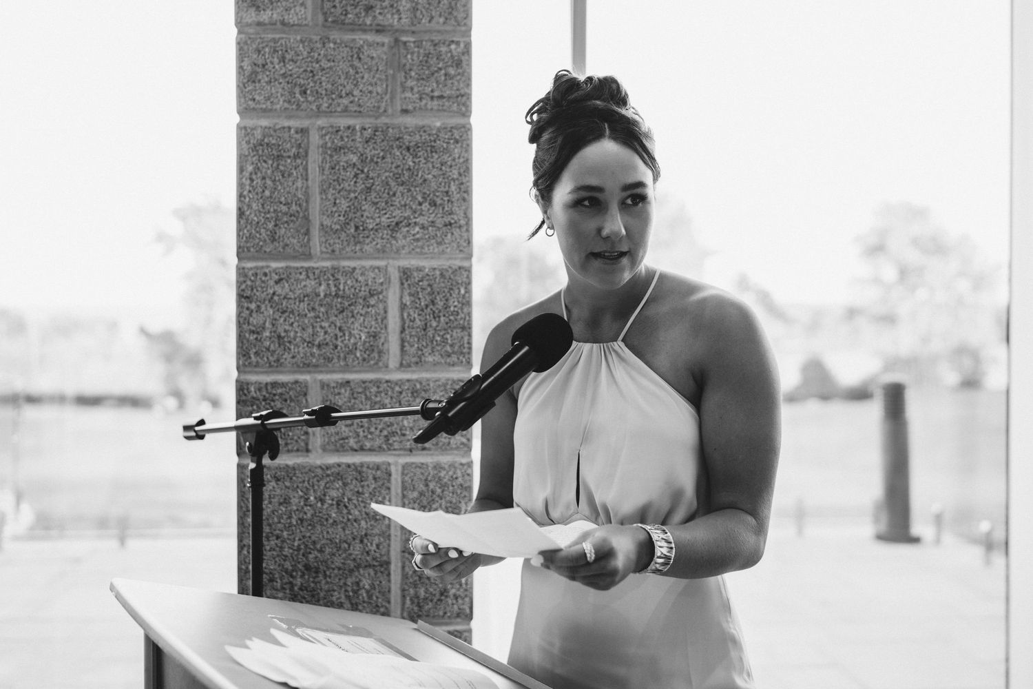 Woman in halter dress speaks at podium with microphone at outdoor event in black and white photo.