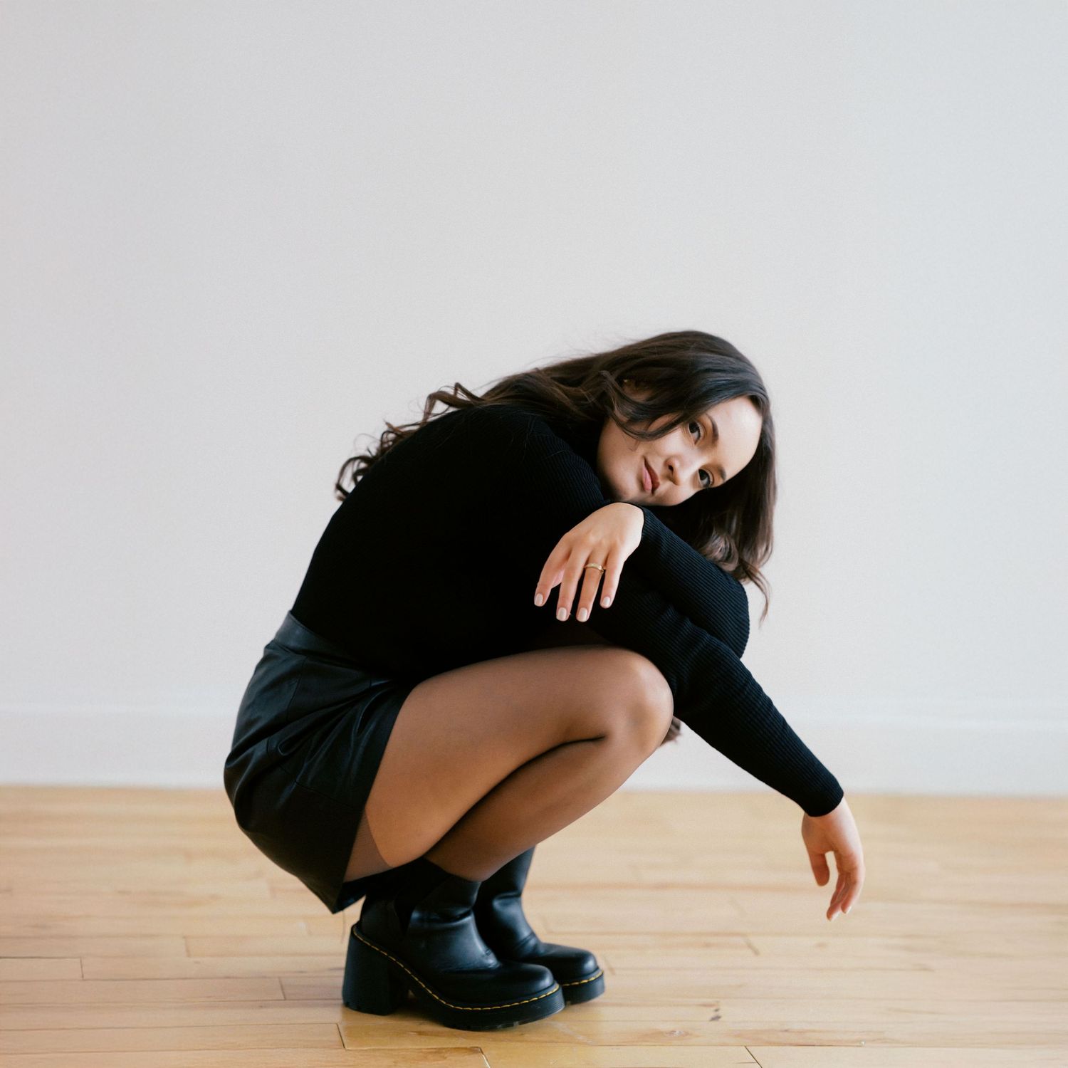 Artistic portrait of someone crouching in a black sweater and combat boots on a wooden floor.