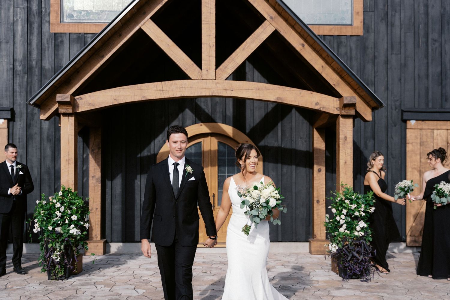 Wedding party poses in front of a rustic black barn with exposed wooden beams and white trim.