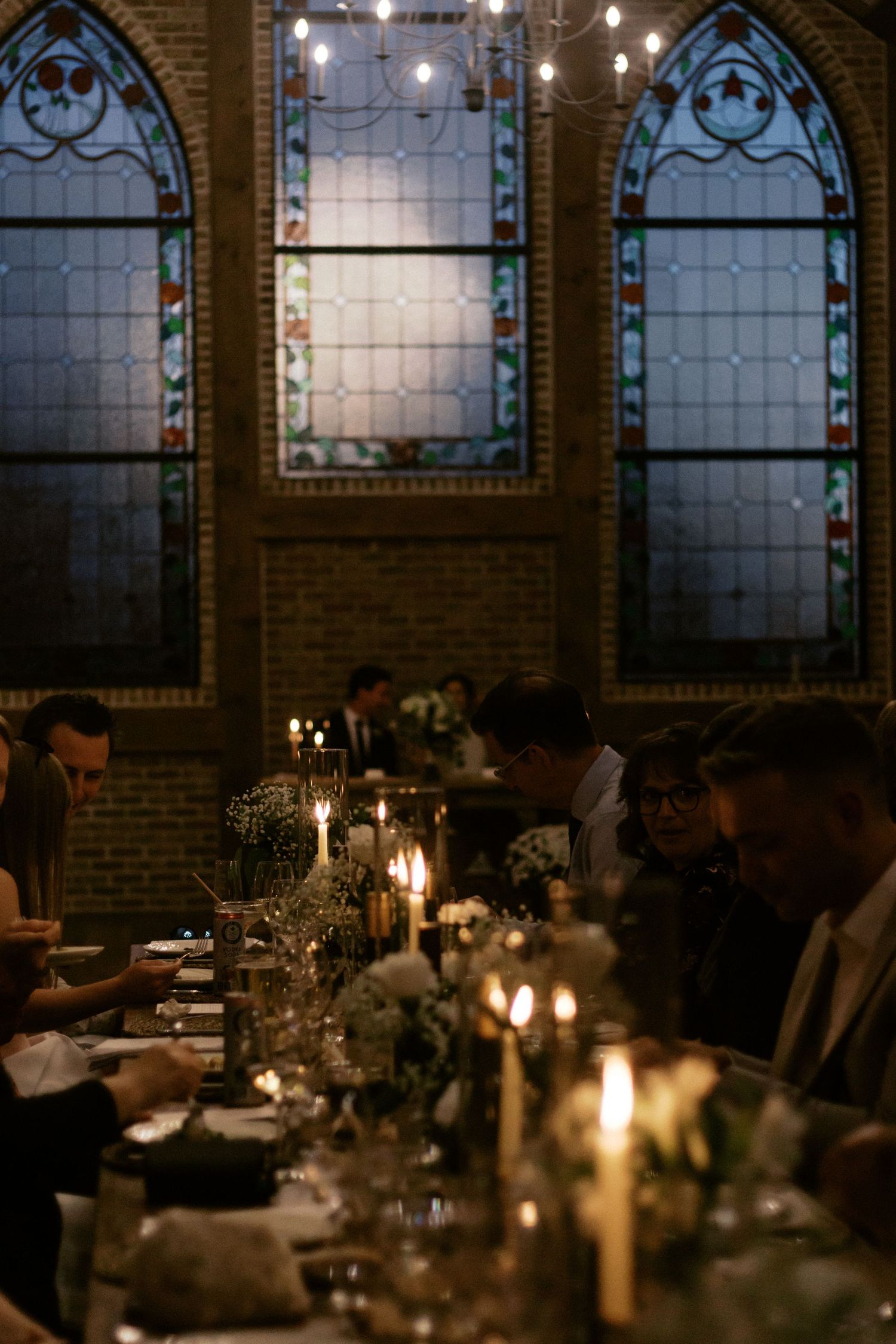 Candlelit dining tables set against ornate stained glass windows in a dimly lit brick-walled restaurant interior.