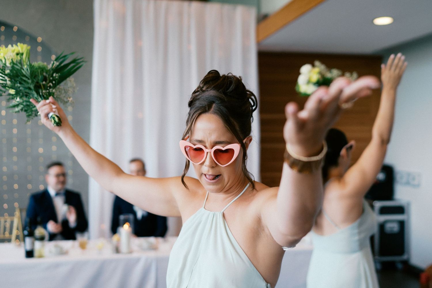 Joyful bridesmaid in heart-shaped sunglasses raises bouquet at wedding reception celebration.