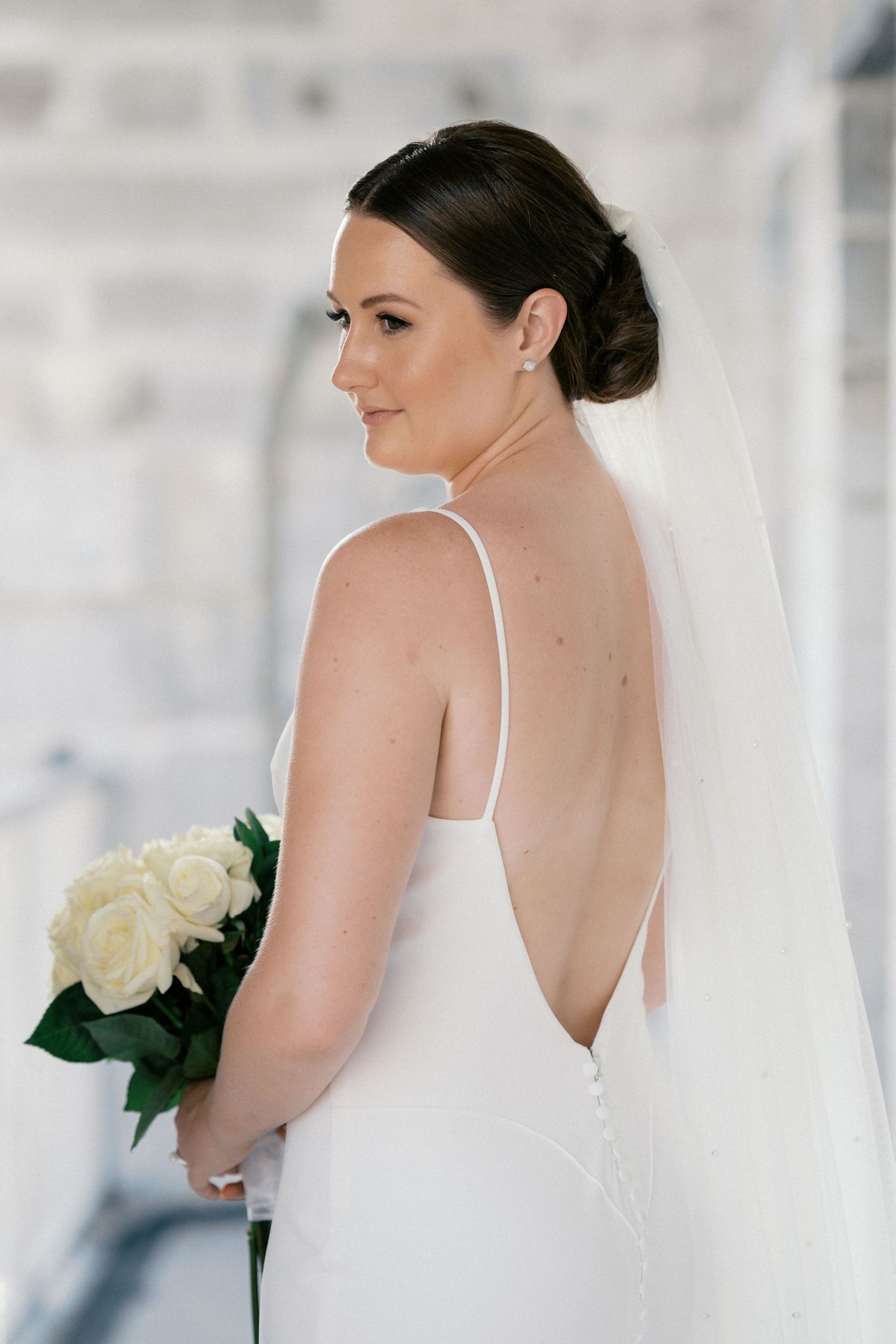 Elegant bride in a backless white gown holding white roses, showcasing her veil and updo hairstyle.