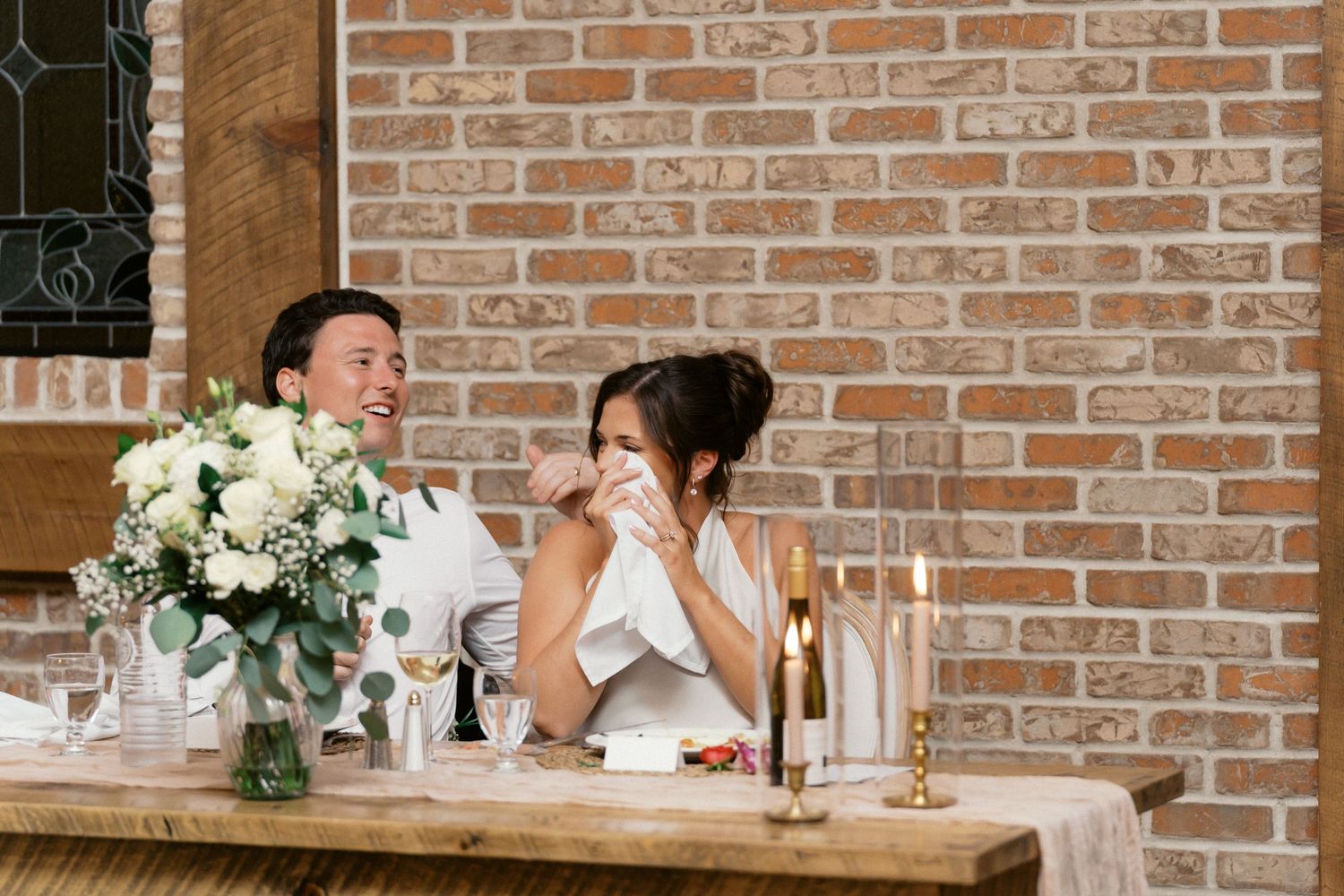 Two people share an emotional moment at a reception table with white floral arrangements against a brick wall.