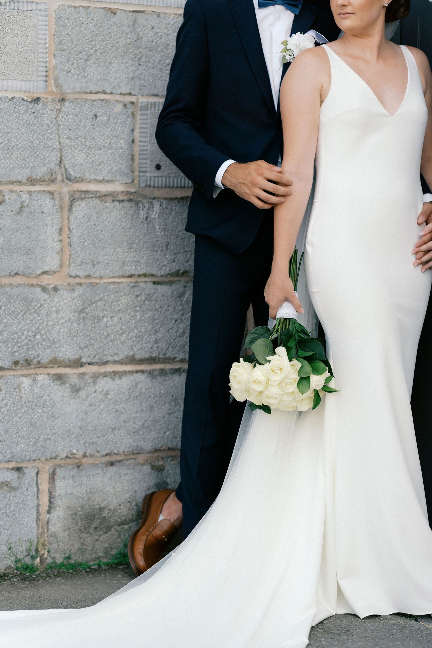 Bride in sleek white gown holding ivory rose bouquet stands beside groom in navy suit against stone wall.