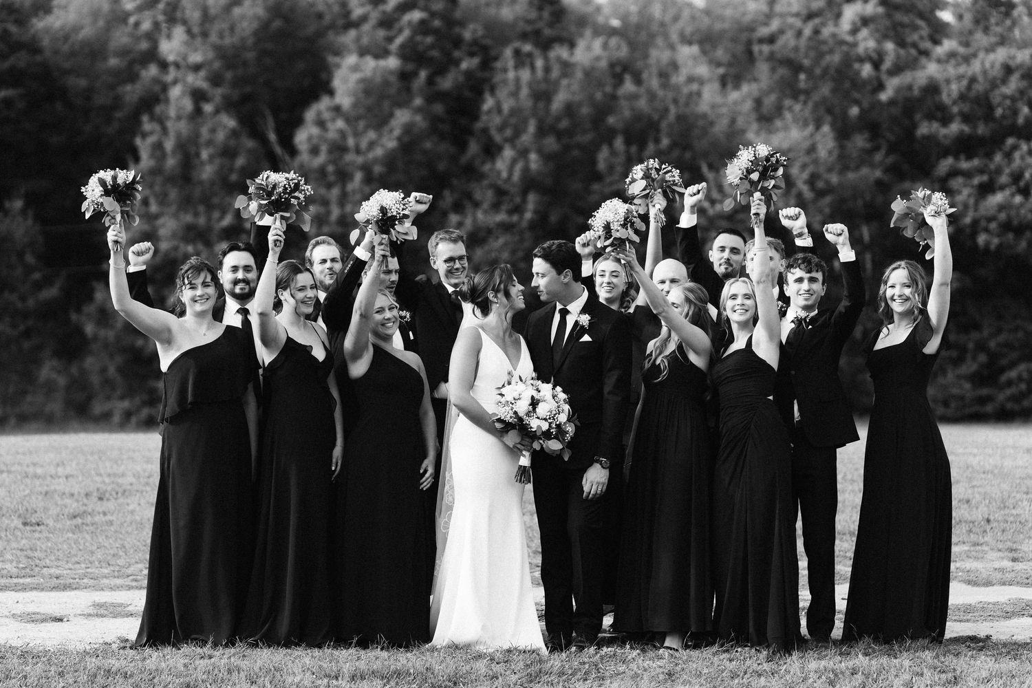 Wedding party in black attire poses together on a sunny outdoor lawn.