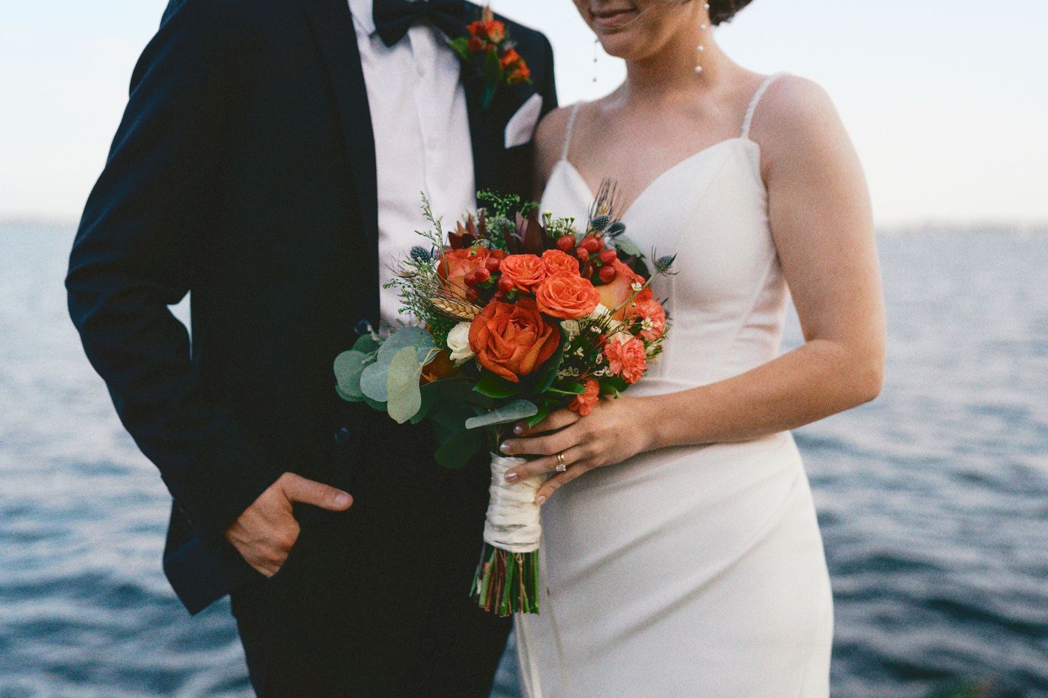 Close-up of newlyweds with coral and eucalyptus bouquet against ocean view.