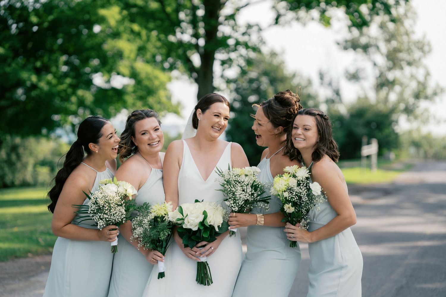 Bride and bridesmaids in white and mint dresses laughing together outdoors holding bouquets.