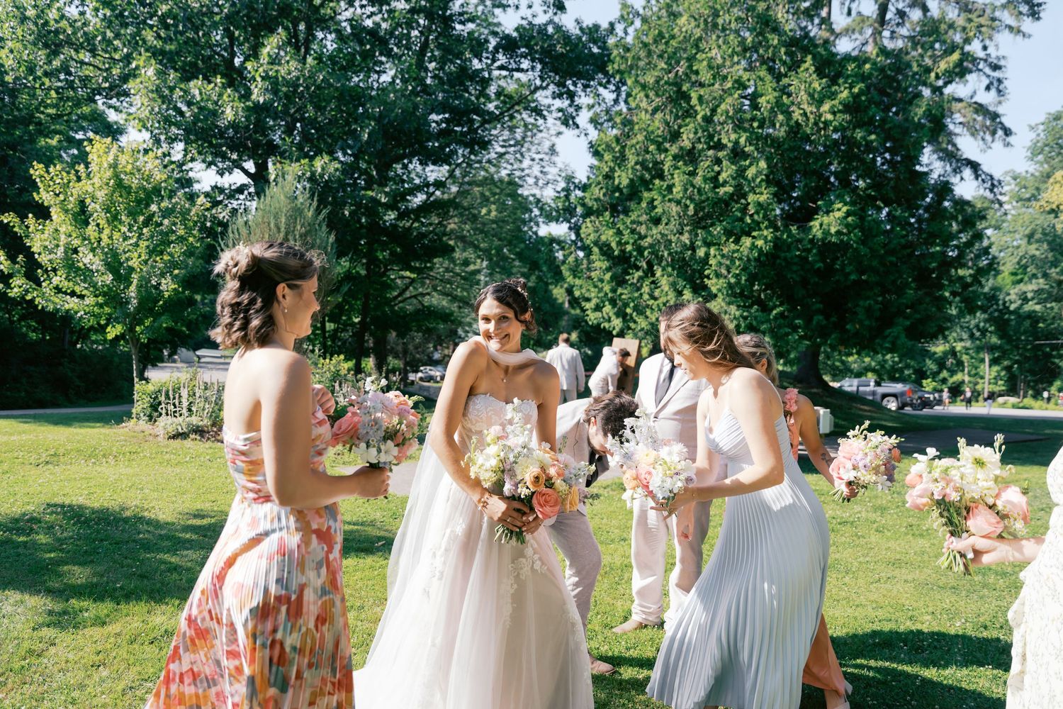 Wedding party poses for photos on a sunny lawn surrounded by trees and natural greenery.