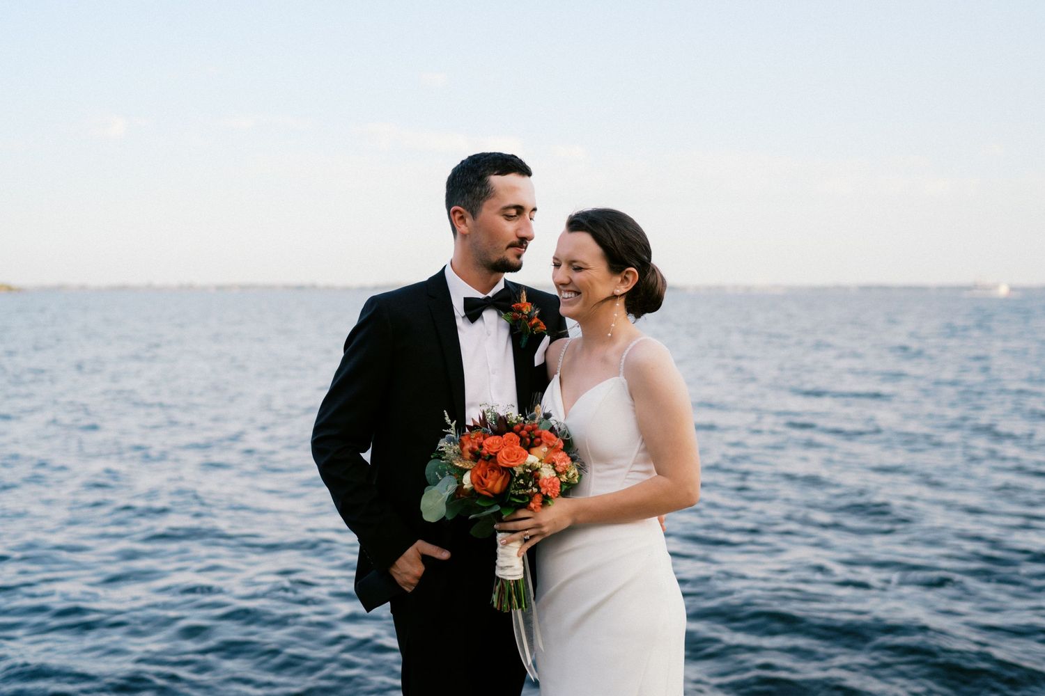 Waterfront wedding portrait with couple holding vibrant orange bouquet.