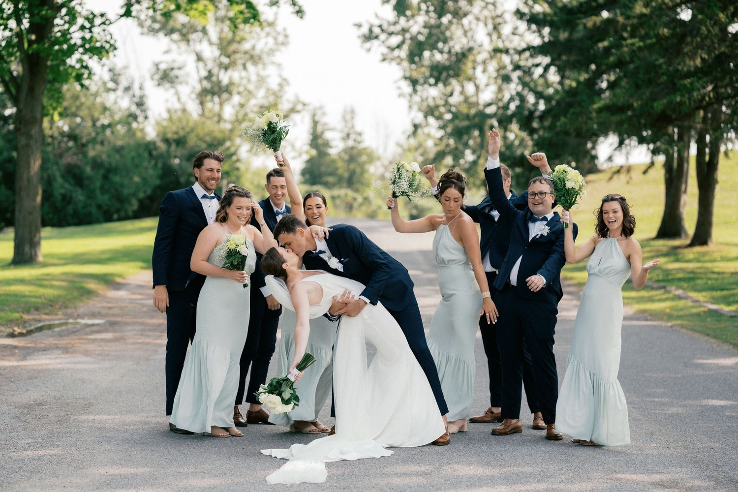 Joyful wedding party celebrating outdoors on tree-lined path, bridesmaids in sage green gowns.