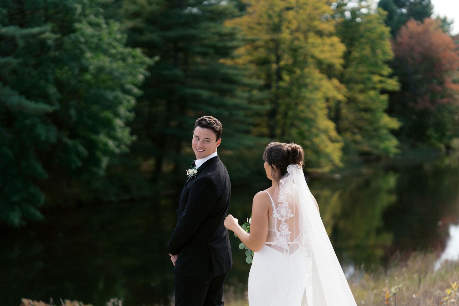 Wedding party gathered outdoors in autumn with trees and lake in background.