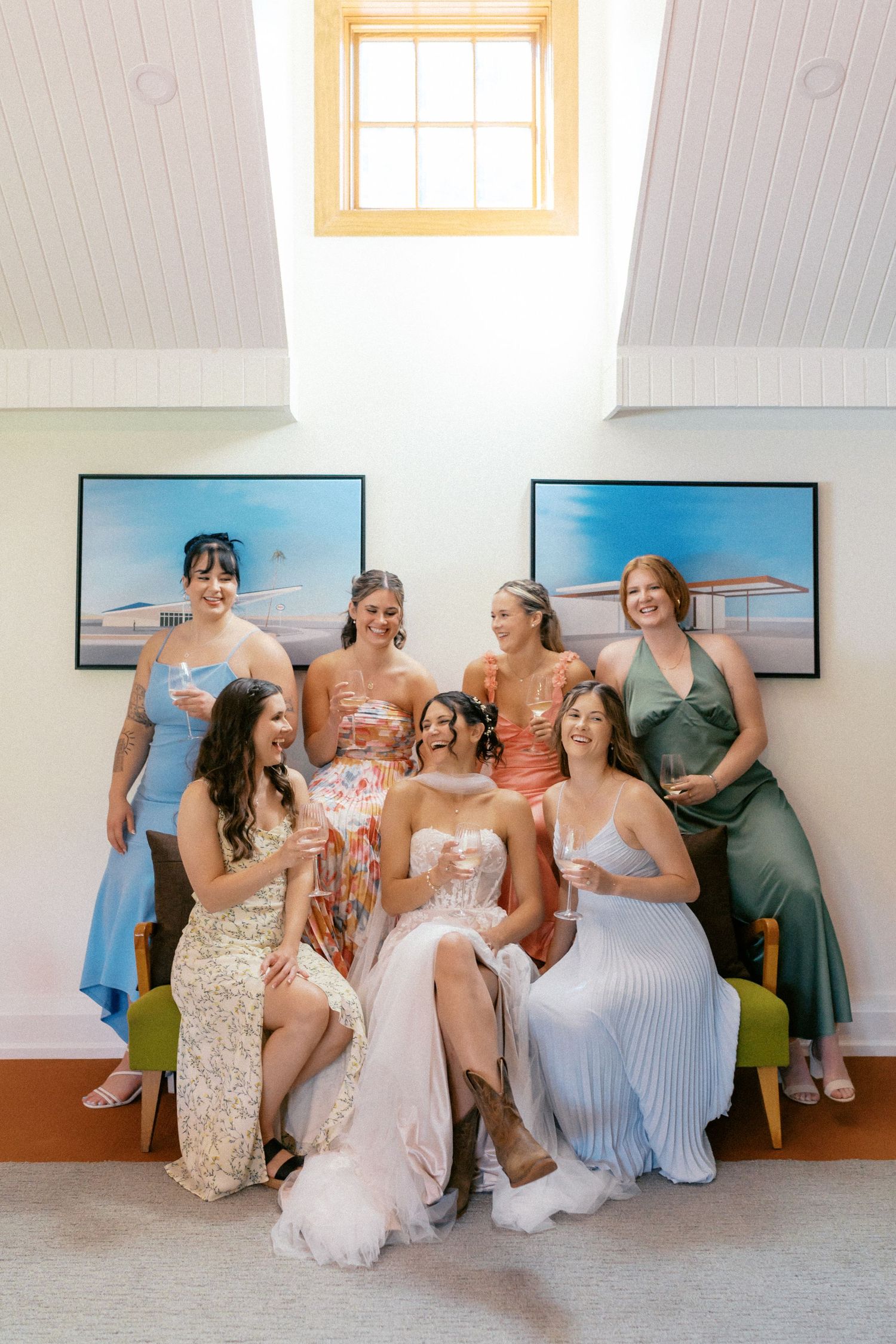 Wedding party poses together on a couch beneath framed artwork in a bright room with a skylight.