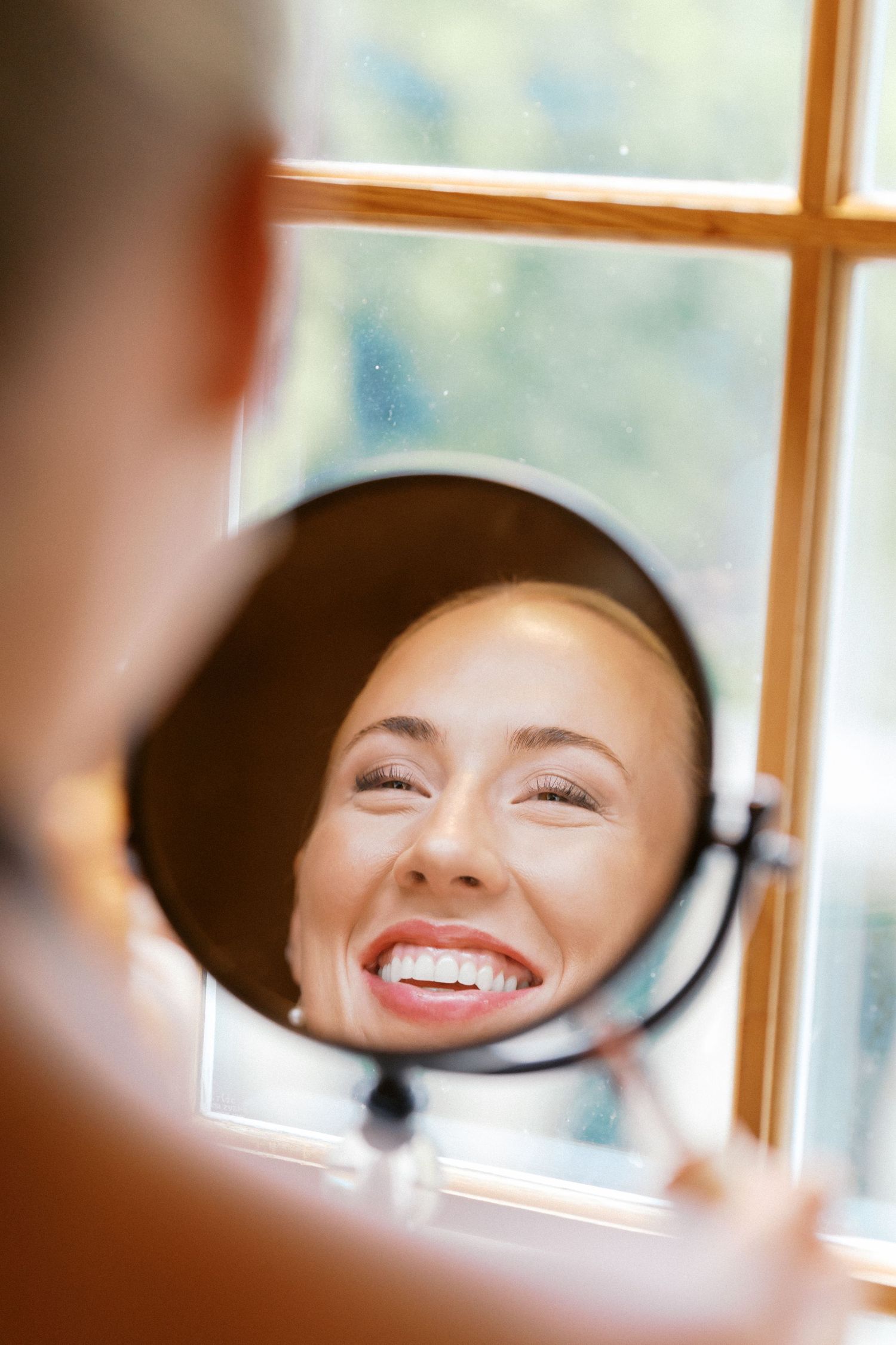 A reflection in a round makeup mirror shows a bright smile near a sunny window.