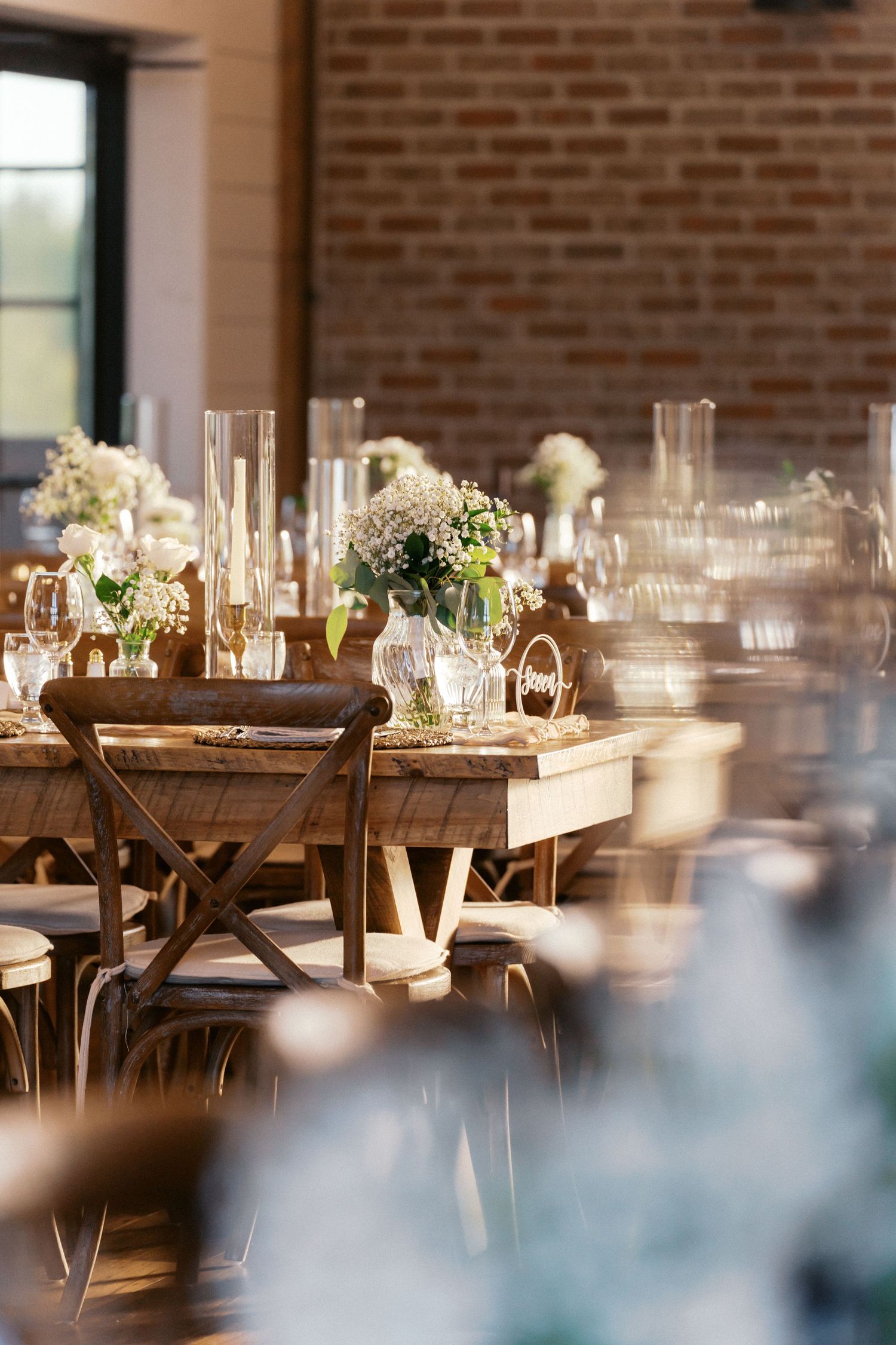 Elegant wedding reception tables set with wooden chairs and floral centerpieces in a brick-walled venue.