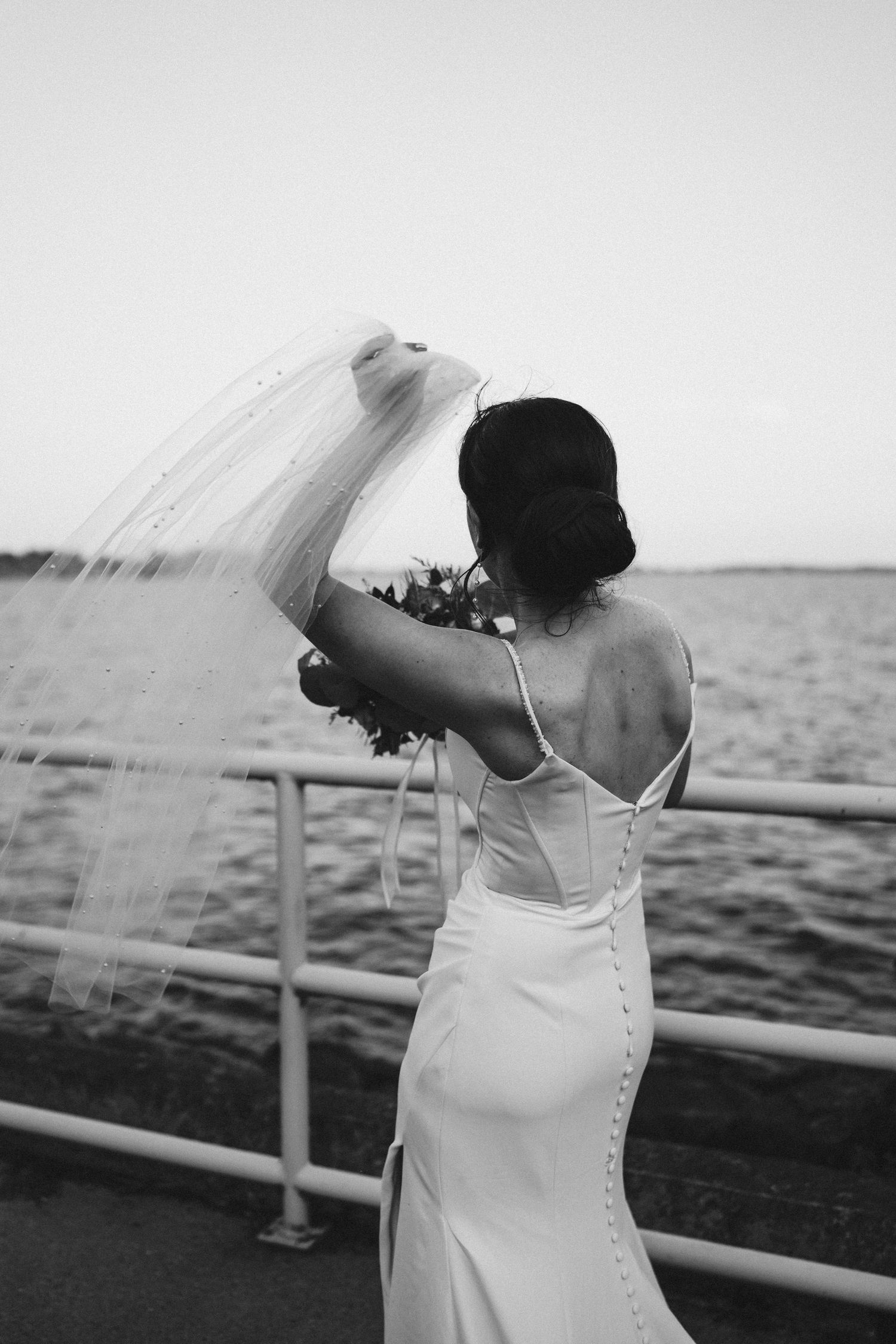 Black and white photo of bride in fitted dress holding veil on boat deck.