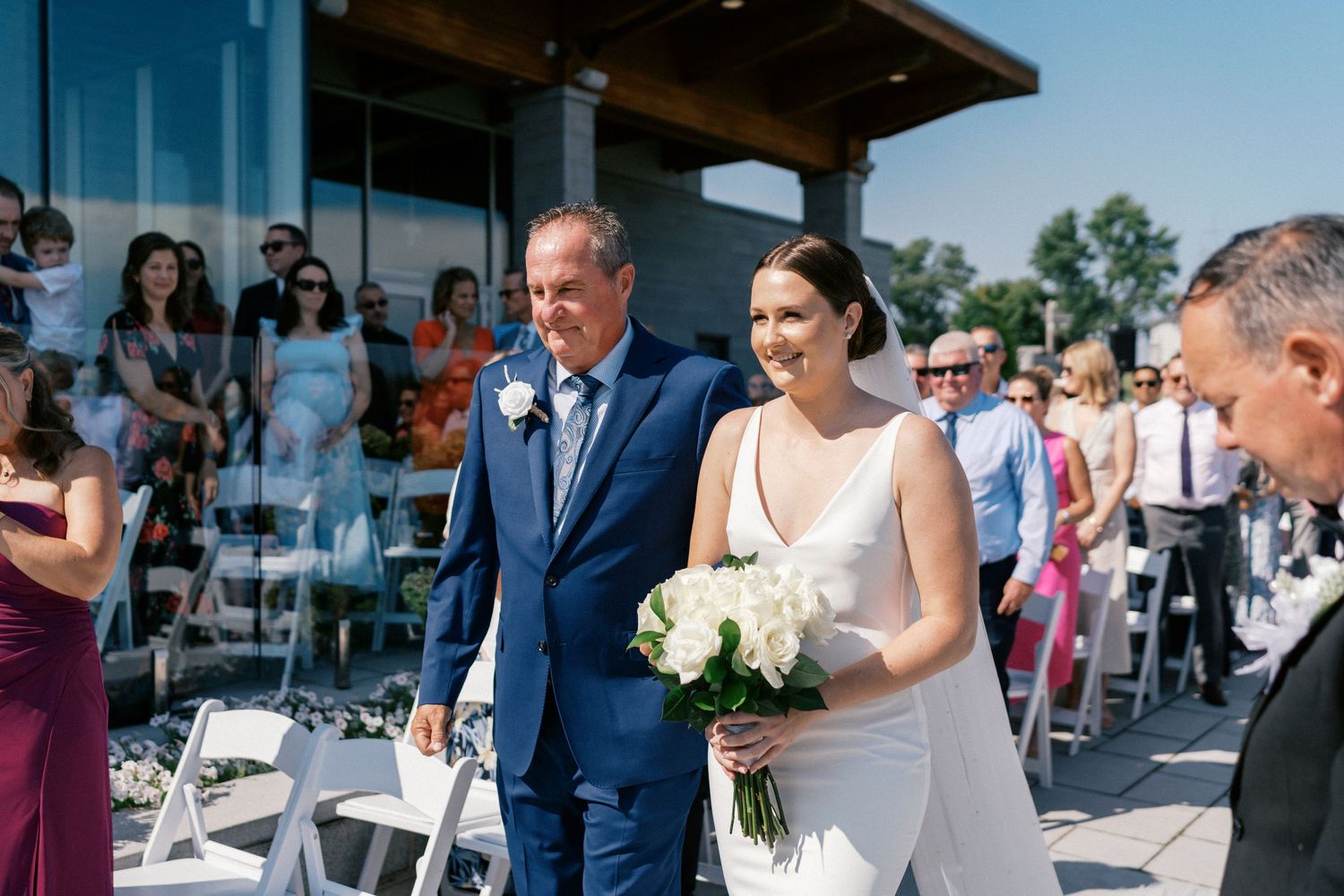 Bride walks down the aisle with escort holding white bouquet at outdoor wedding ceremony.