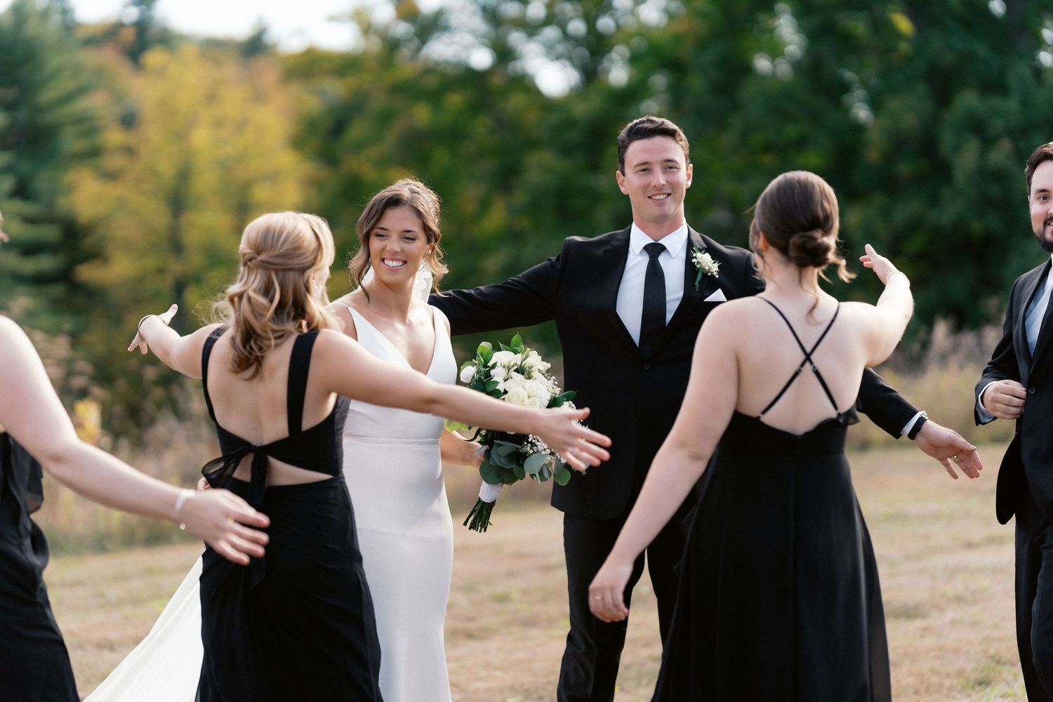 Wedding party in black and white attire celebrating on grassy hill during autumn.