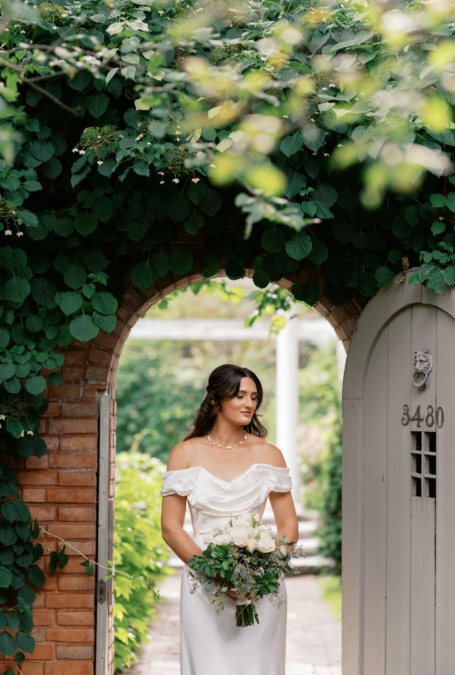 A bride in an off-shoulder white wedding dress holds a bouquet while standing under a vine-covered brick archway.