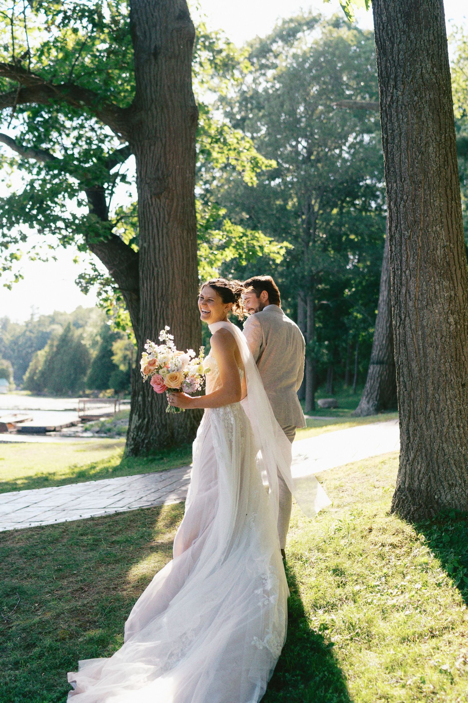 Couple in wedding attire walking together among tall trees in golden sunlight.