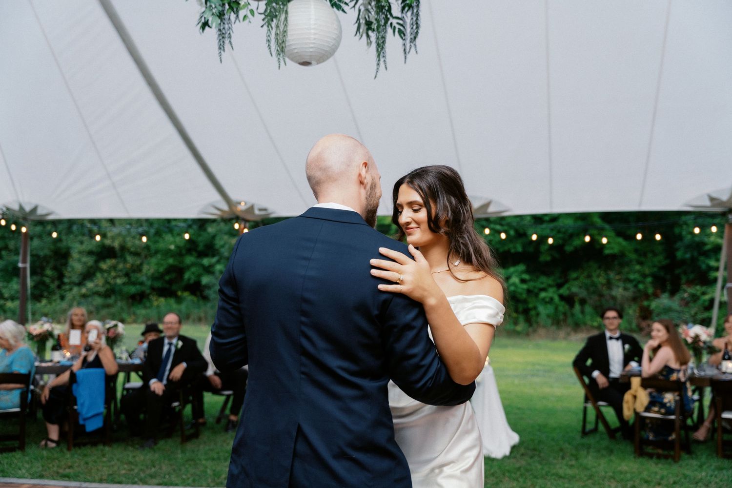 A couple shares their first dance under a white tent at an outdoor wedding reception.