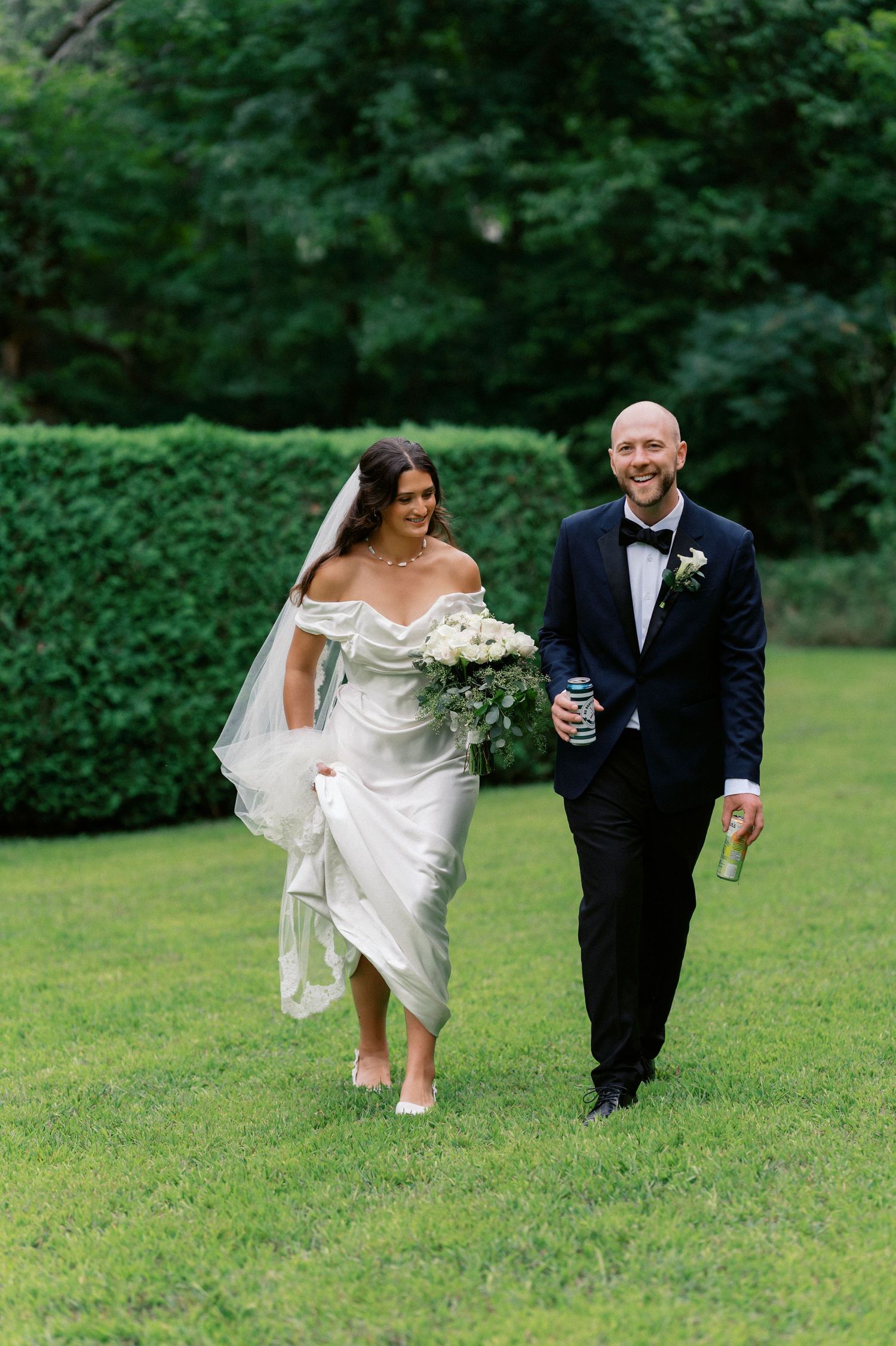 A couple in wedding attire playfully runs across a lush green lawn during their outdoor celebration.