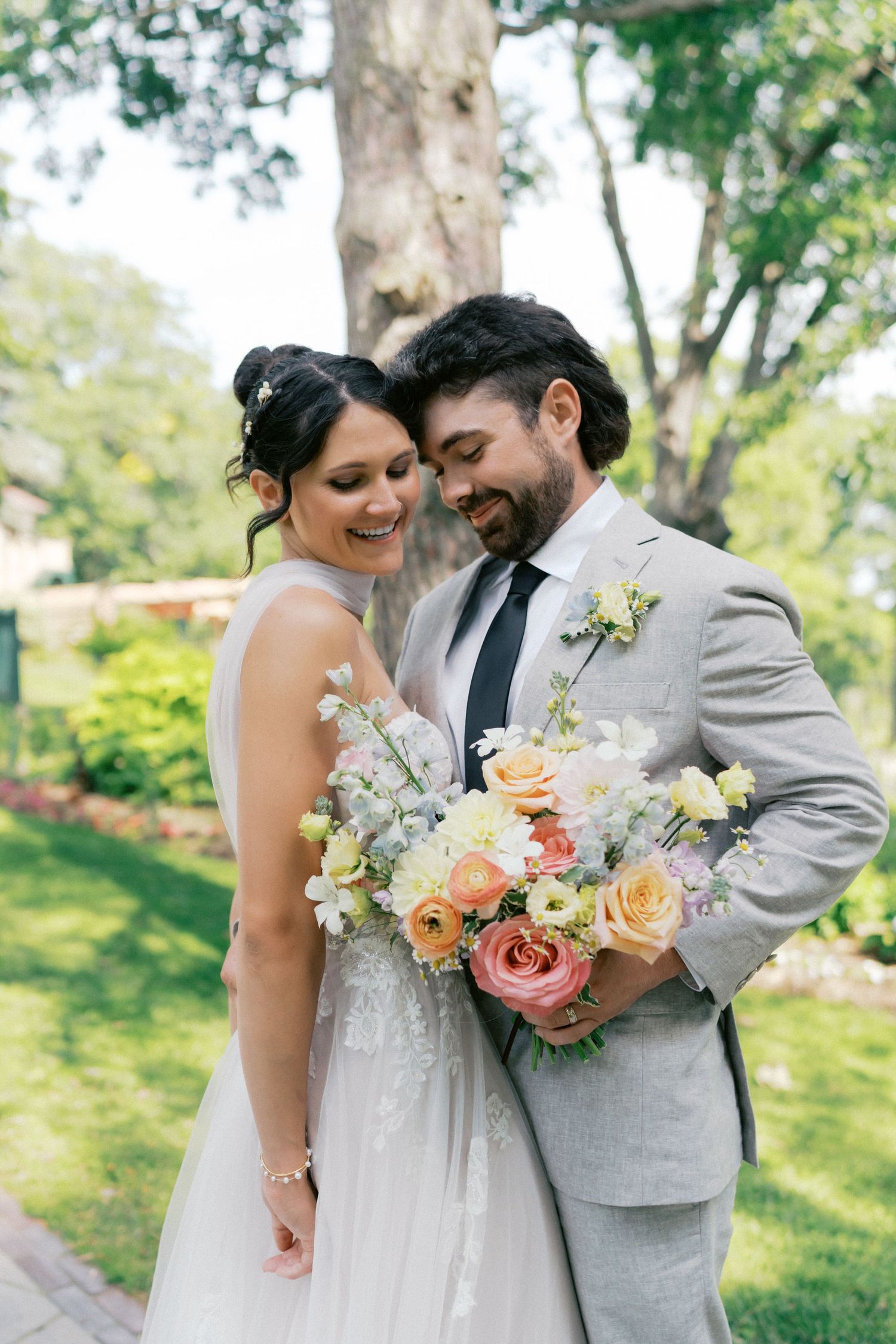 Romantic wedding couple poses with peach and cream floral bouquet in a sunlit garden setting.