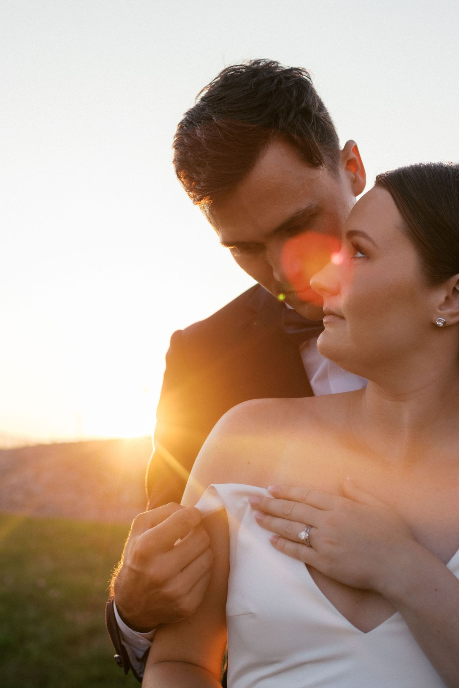 A groom kisses his bride on the cheek at golden hour sunset, her diamond ring visible as they embrace outdoors.