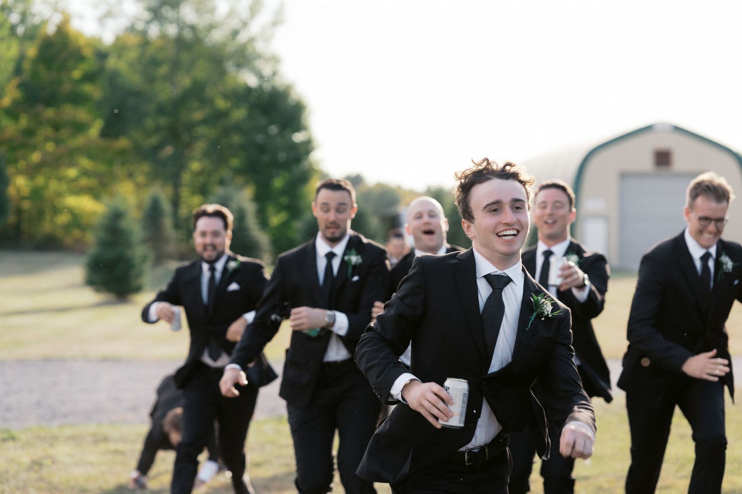 Groomsmen in black suits run and laugh together outdoors near a barn.