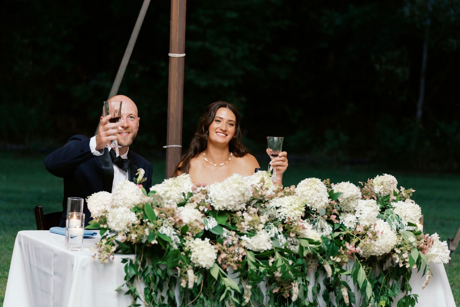 Elegant white floral arrangements and greenery decorate wedding reception head table.