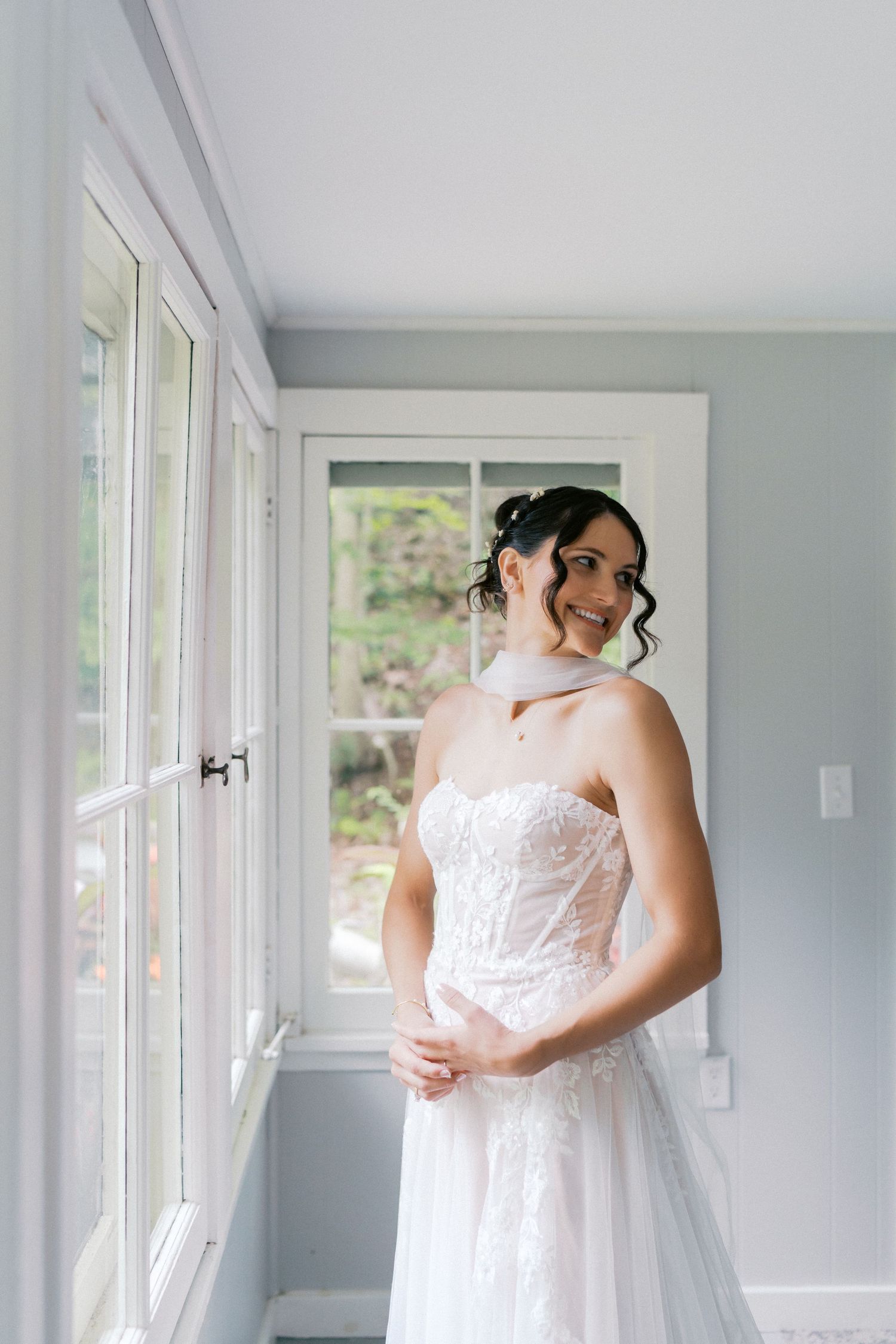 A bride in a white lace wedding dress poses by sunlit windows in a bright room.