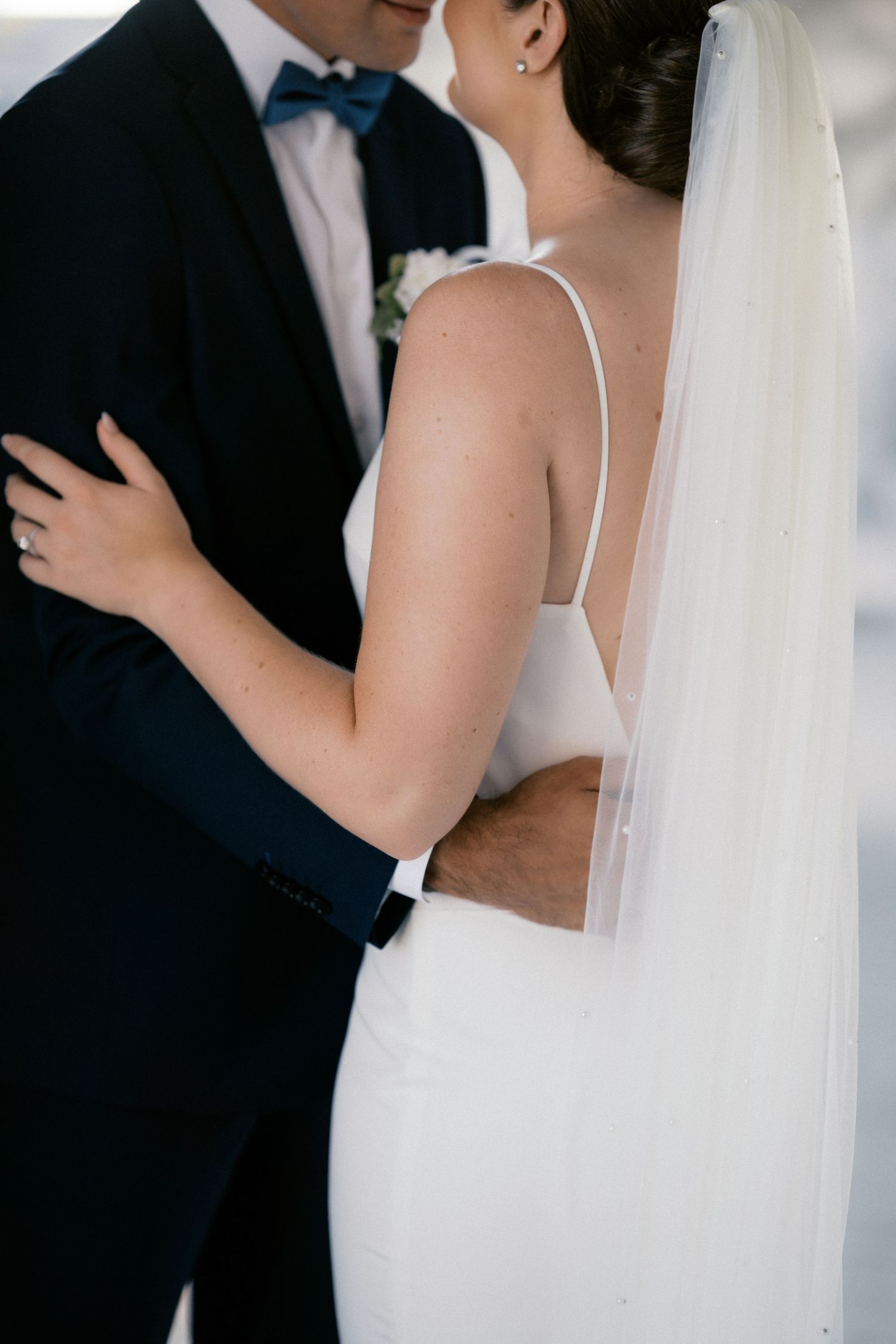 Bride in backless white gown with veil embracing groom in navy suit during romantic wedding portrait.