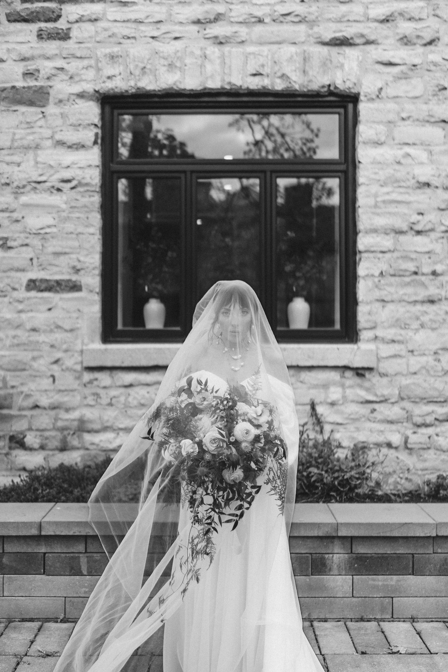 Black and white image of bride from behind with flowing veil and bouquet near brick wall.
