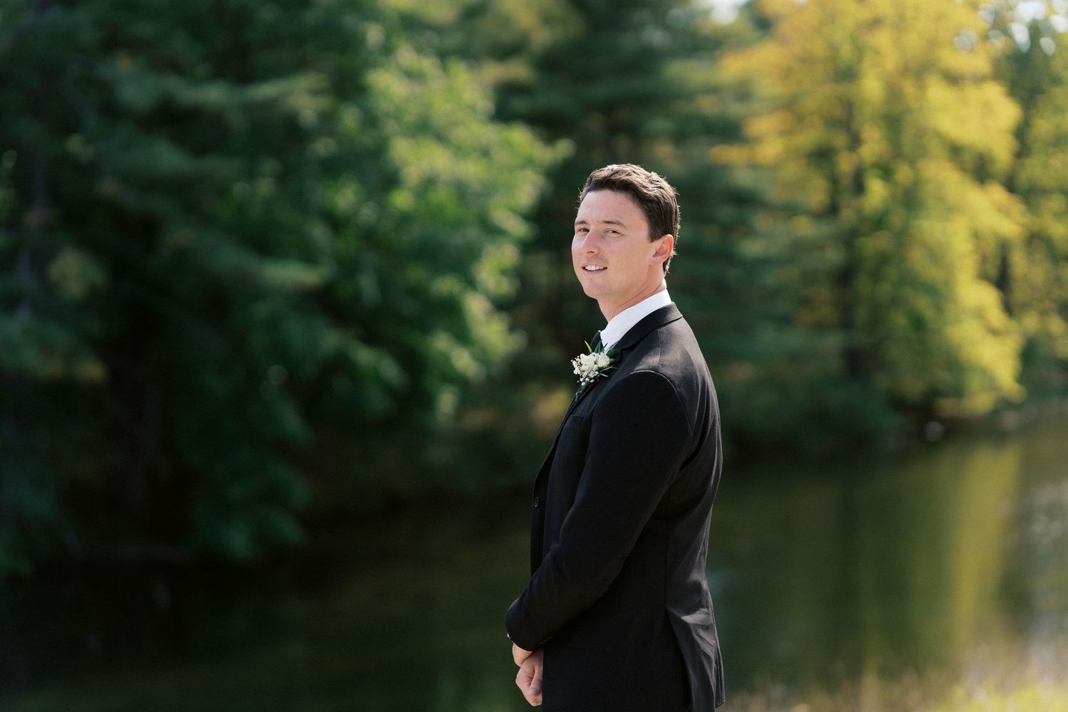 Wedding party members in formal attire standing by a scenic lake with autumn foliage.