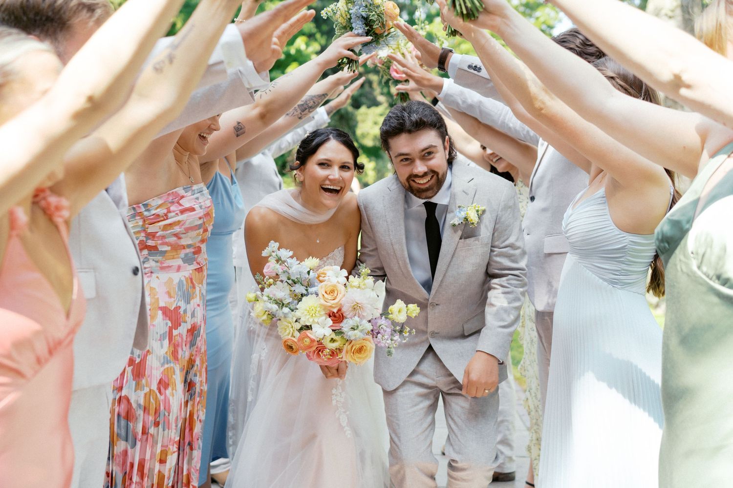 Wedding party celebrates with raised arms forming a tunnel as newlyweds walk through with joyful expressions.
