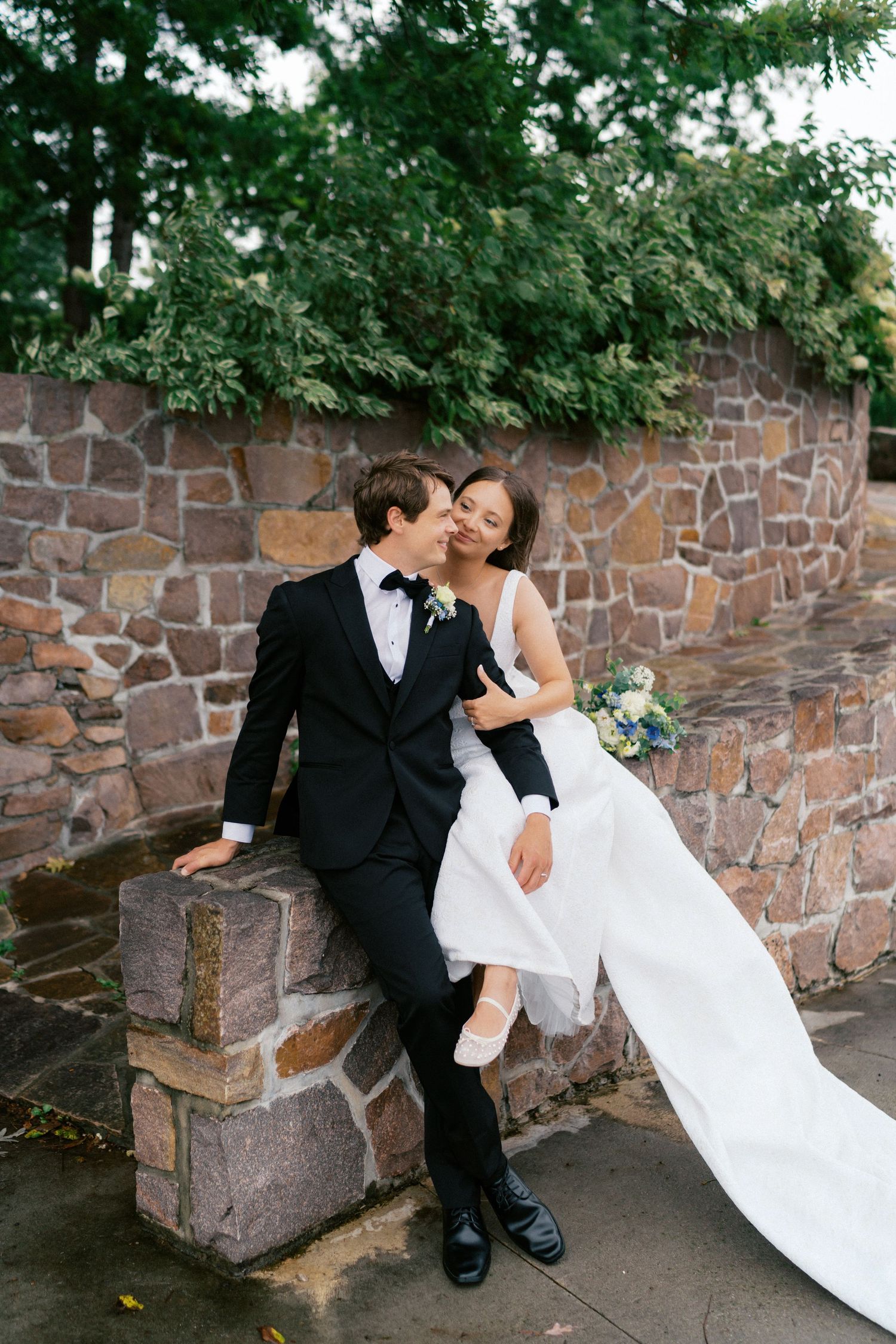 Newlyweds share a romantic moment against a stone wall outdoors.