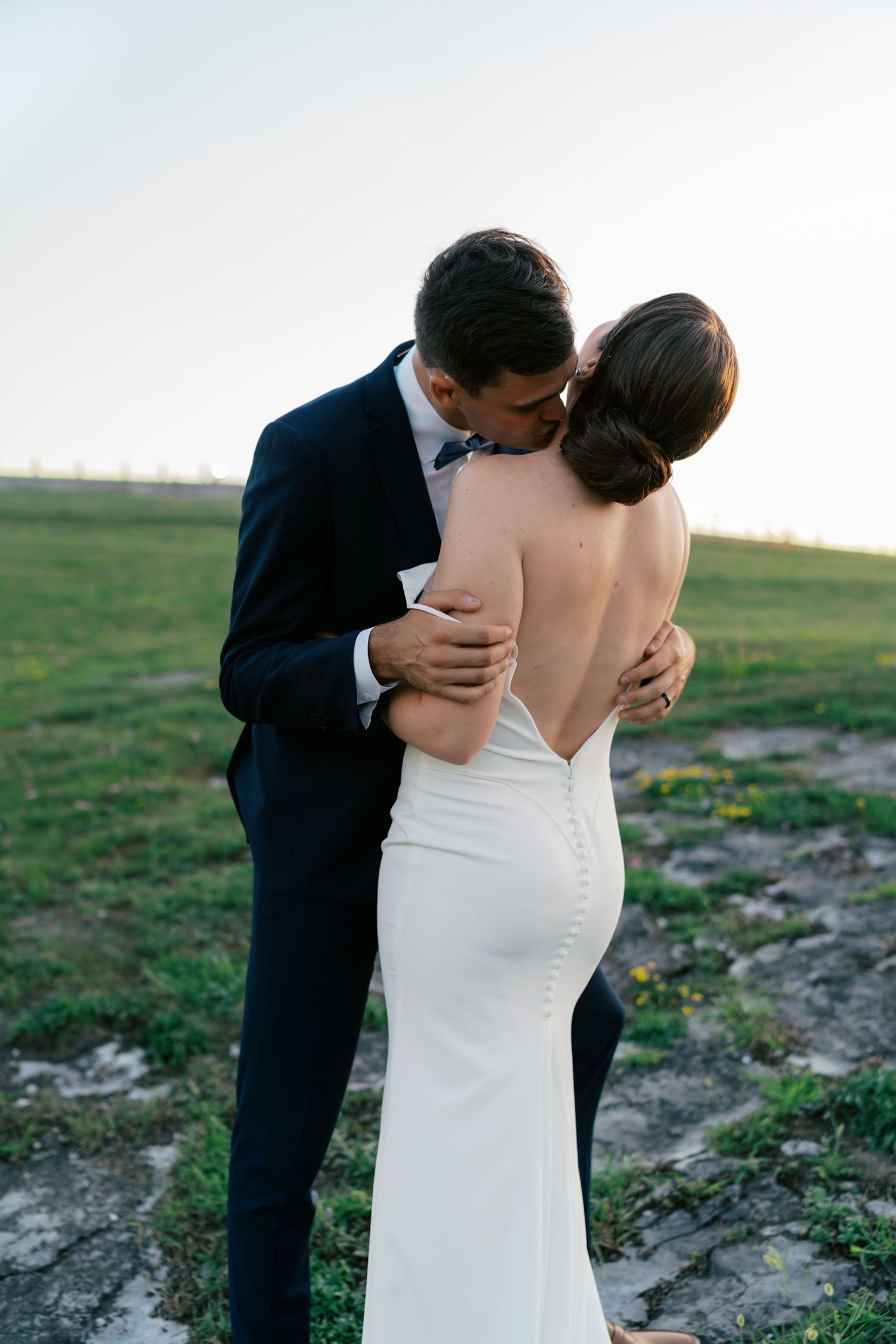 Groom in black tuxedo kissing bride in backless white gown outdoors at sunset wedding.