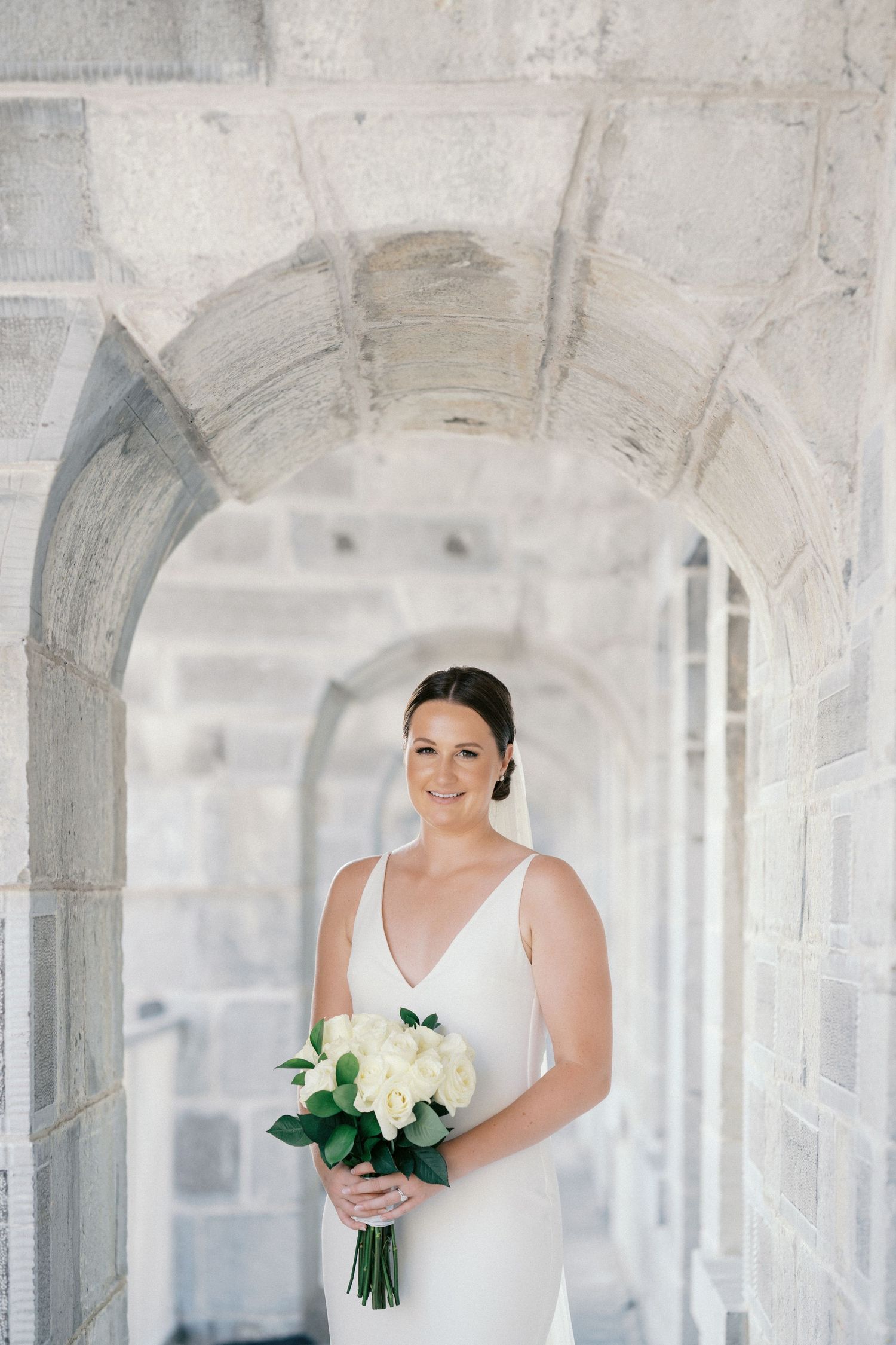 Elegant bride in white gown holding white rose bouquet standing under stone arches in a stunning architectural setting.
