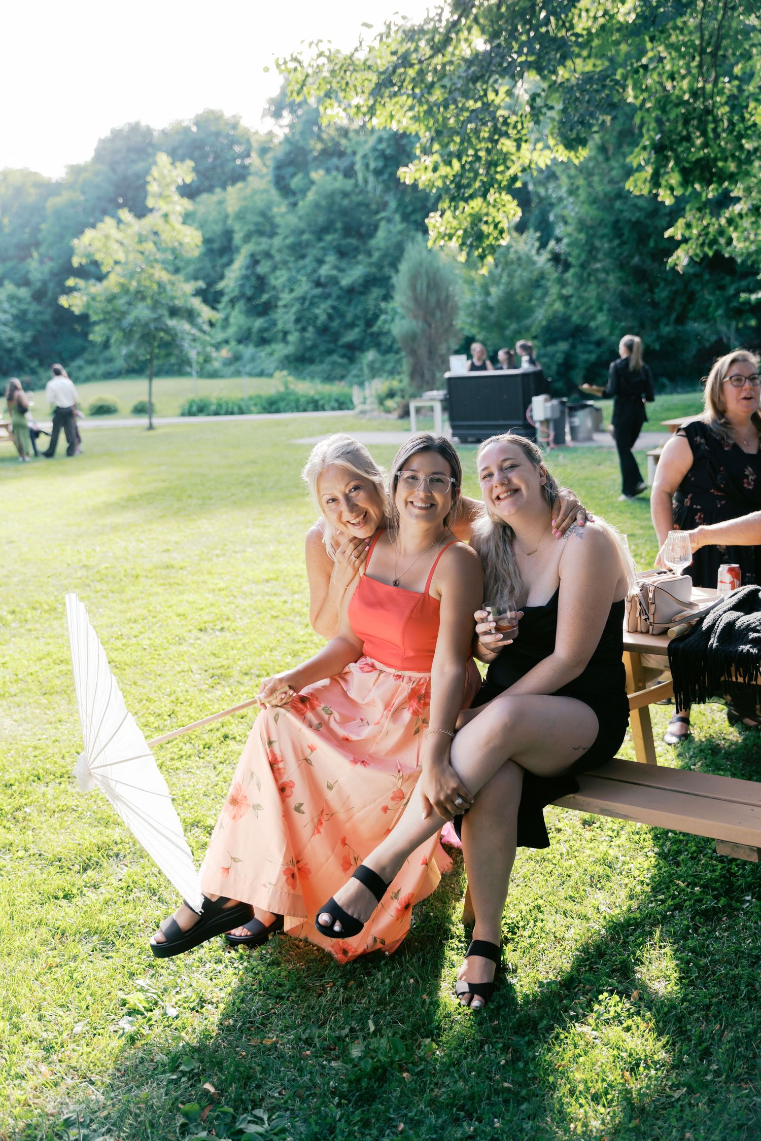 Bridesmaids in coral dresses relax and laugh together on wooden benches in a park setting.