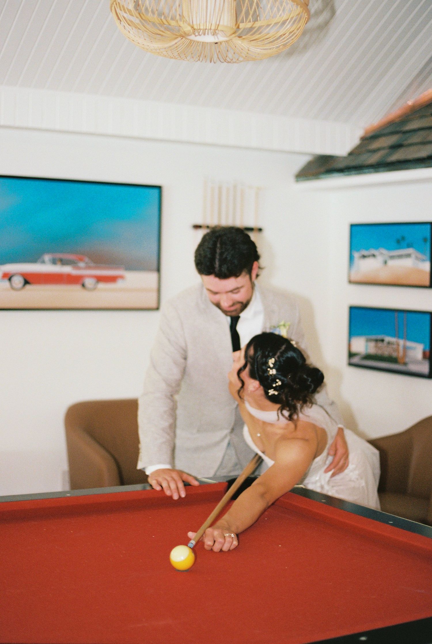 Newlyweds share a playful moment at a pool table in a room decorated with vintage car artwork.