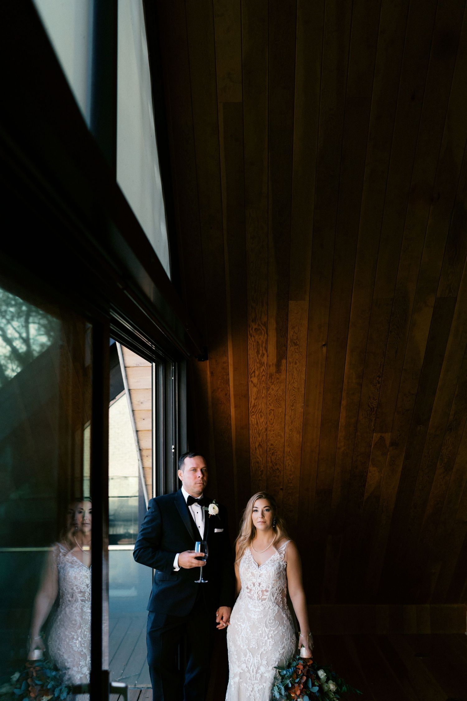 Bride and groom in formal attire pose by window with dramatic lighting.