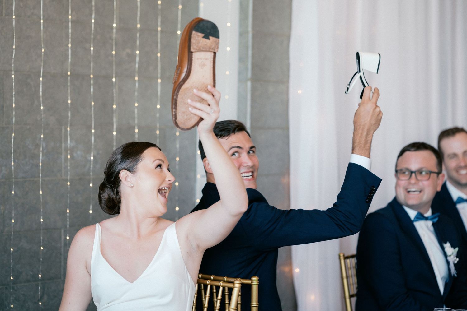 Bride and groom laughing while holding up shoes during a fun wedding reception game at elegantly decorated venue.