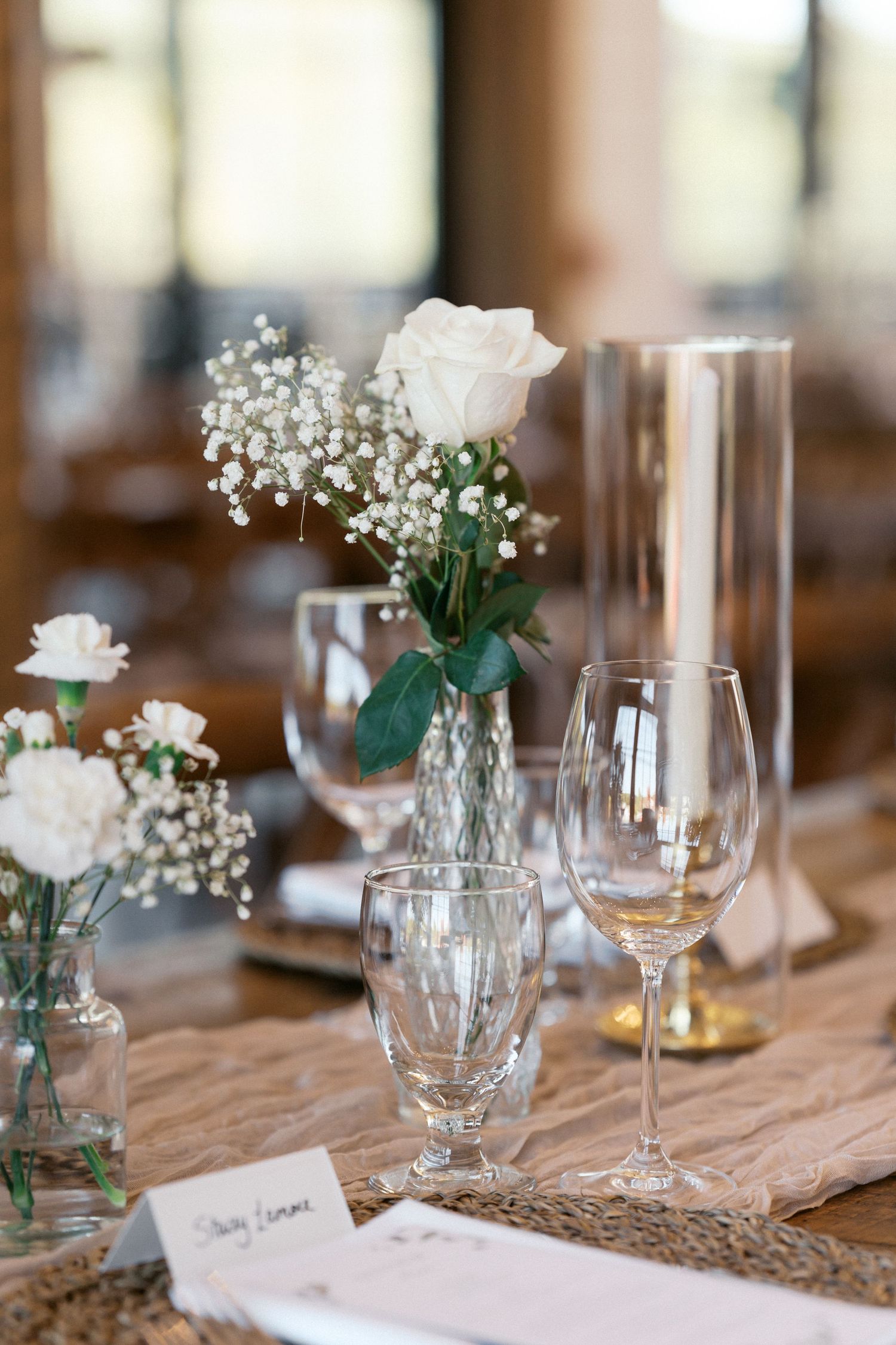 Elegant rustic table setting with wine glasses and small white flower arrangements in mason jars on wooden surface.