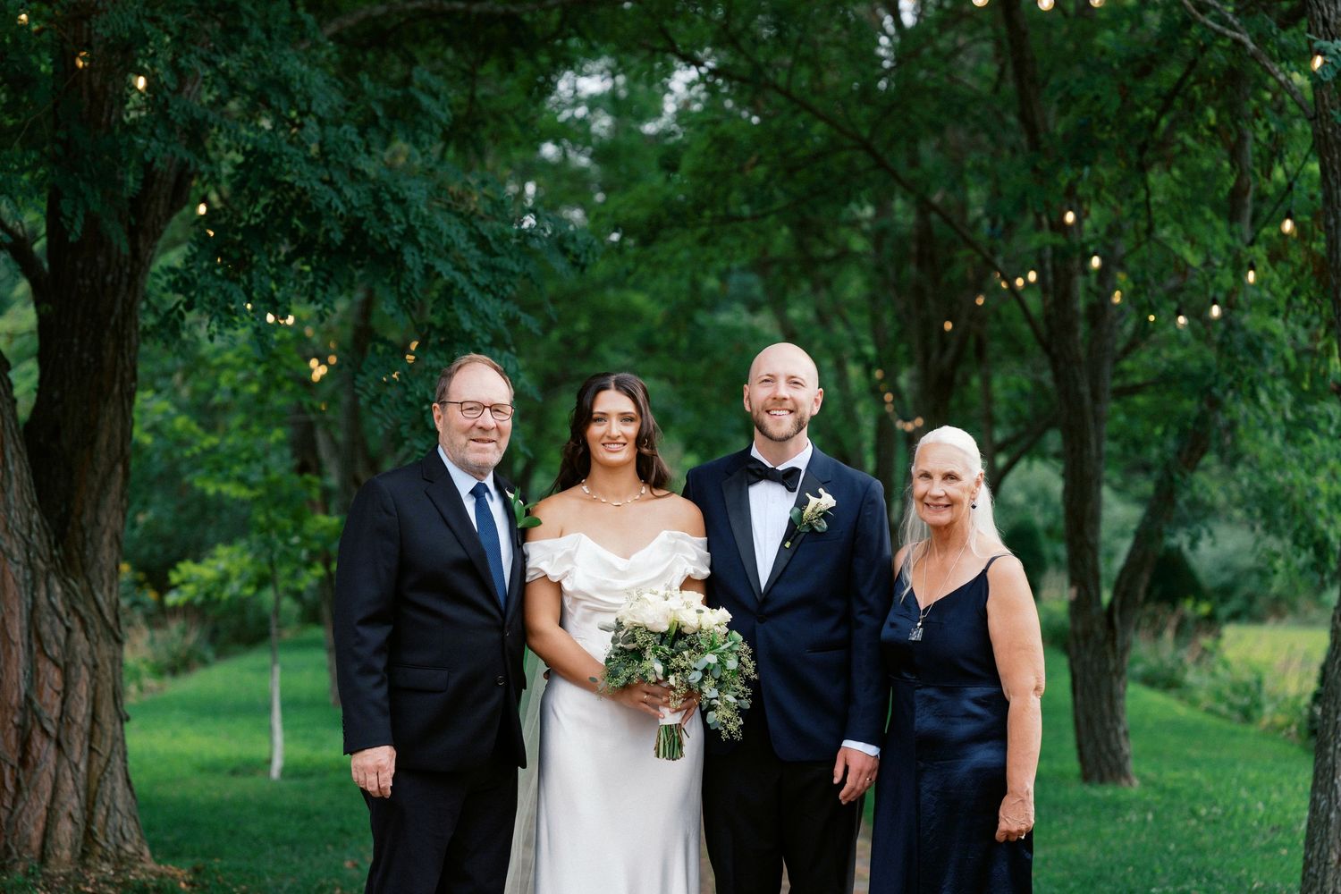 Wedding party poses together outdoors under trees at an elegant garden ceremony.
