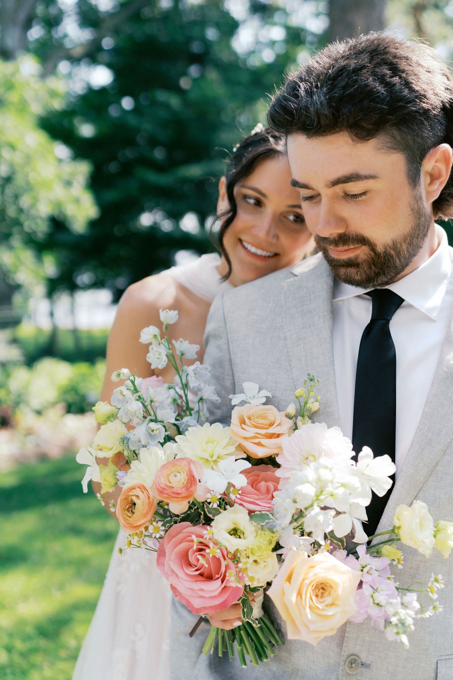 A romantic wedding couple embraces outdoors while holding a beautiful bouquet of pink and peach roses.