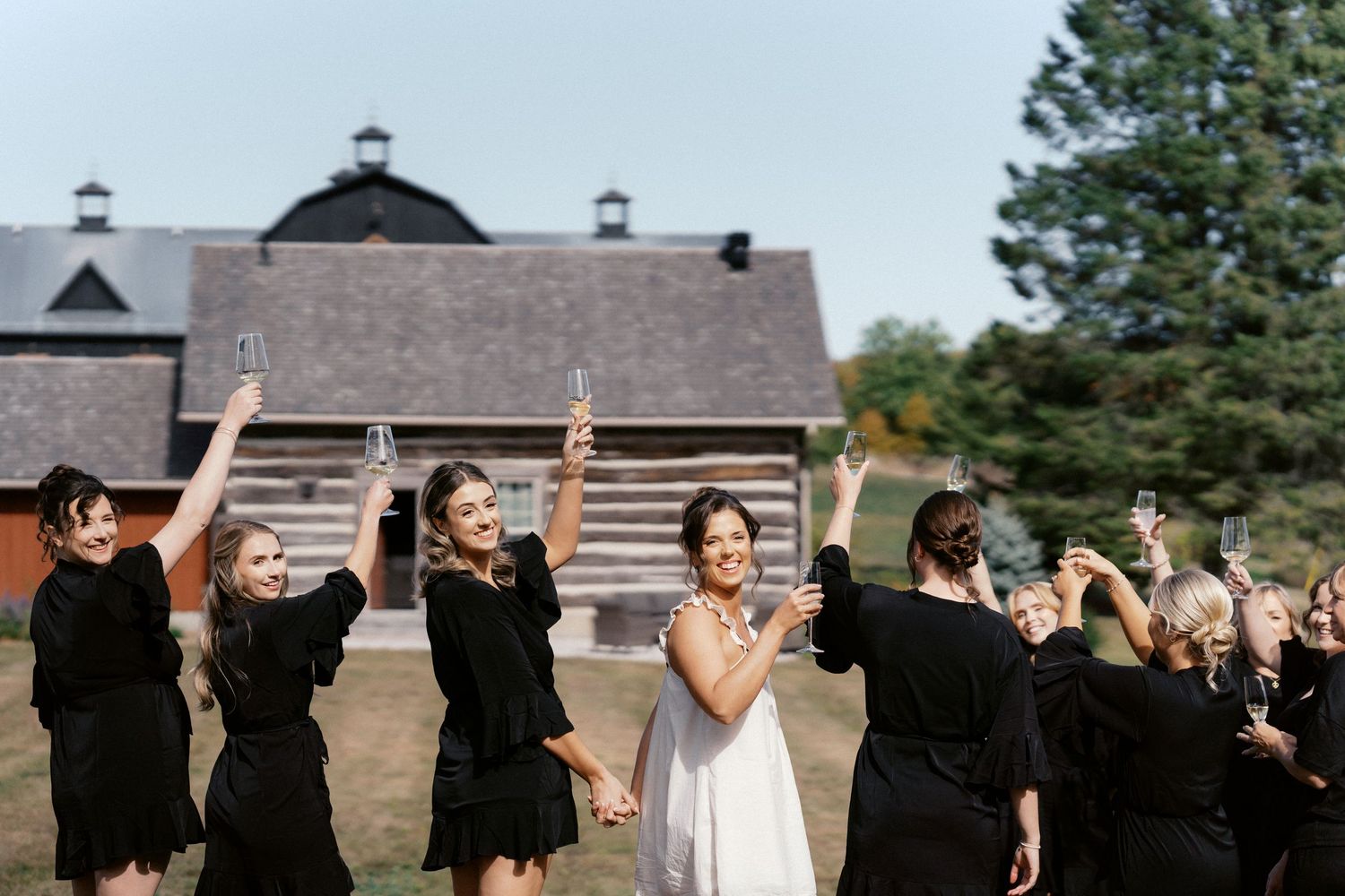 Wedding party in black dresses celebrating outdoors with joyful expressions near a barn.
