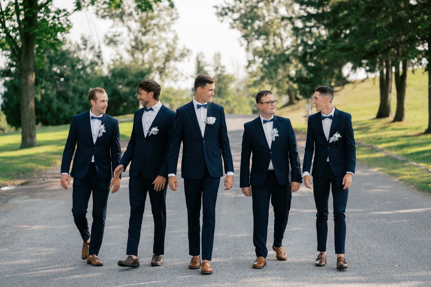 Five groomsmen in navy suits and bow ties walk together on a tree-lined path at an outdoor wedding.
