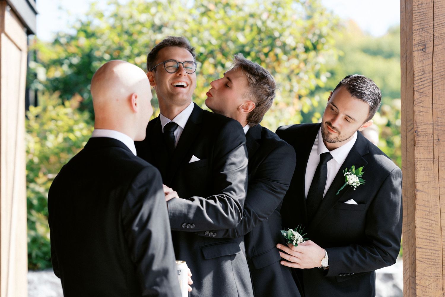 Groomsmen in black suits share emotional hugs and laughter during outdoor wedding celebrations.