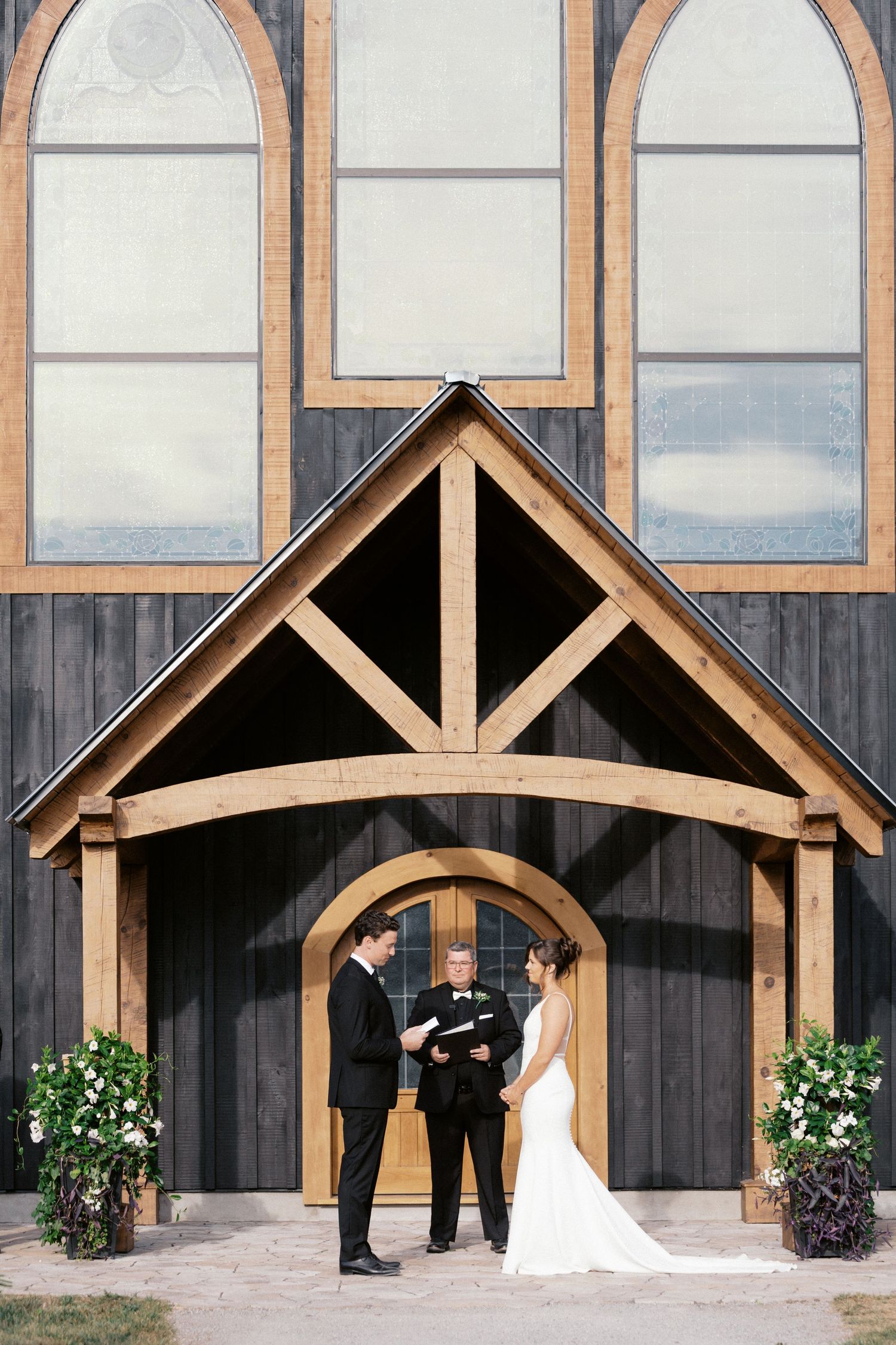 Wedding ceremony taking place under a rustic wooden archway entrance with tall arched windows and floral arrangements.