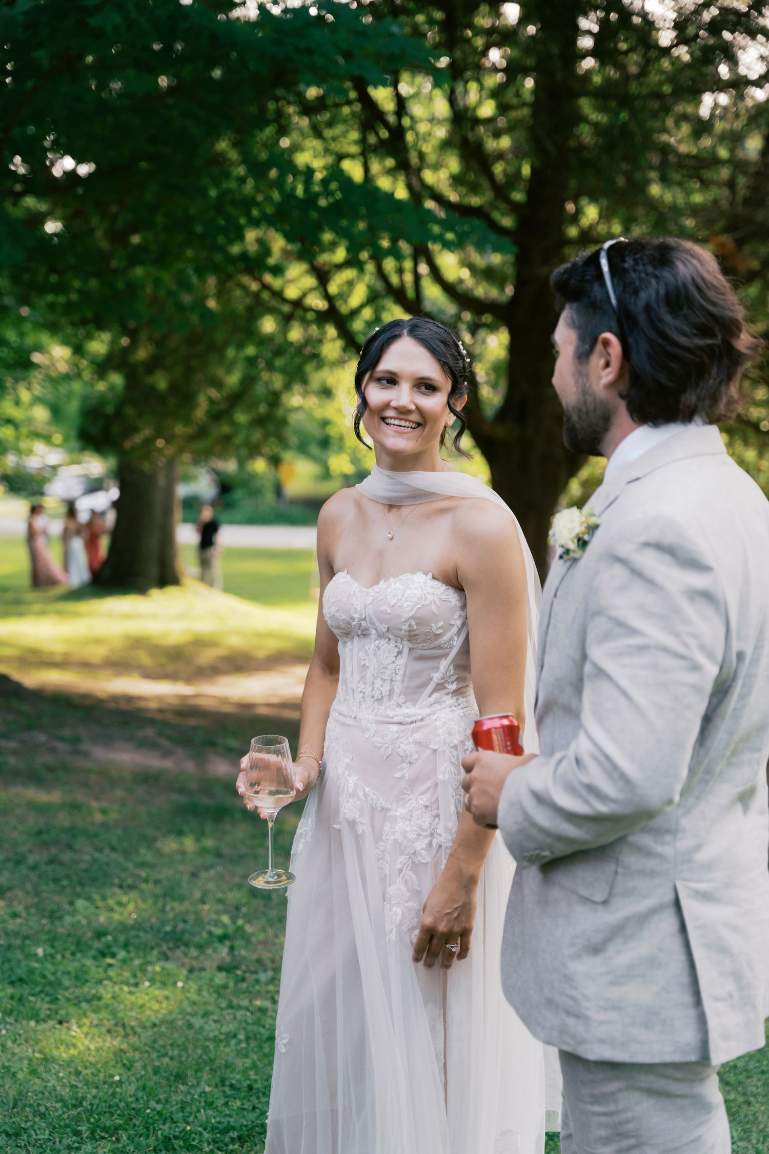 Wedding couple shares intimate moment while holding drinks on grassy lawn under tree canopy.